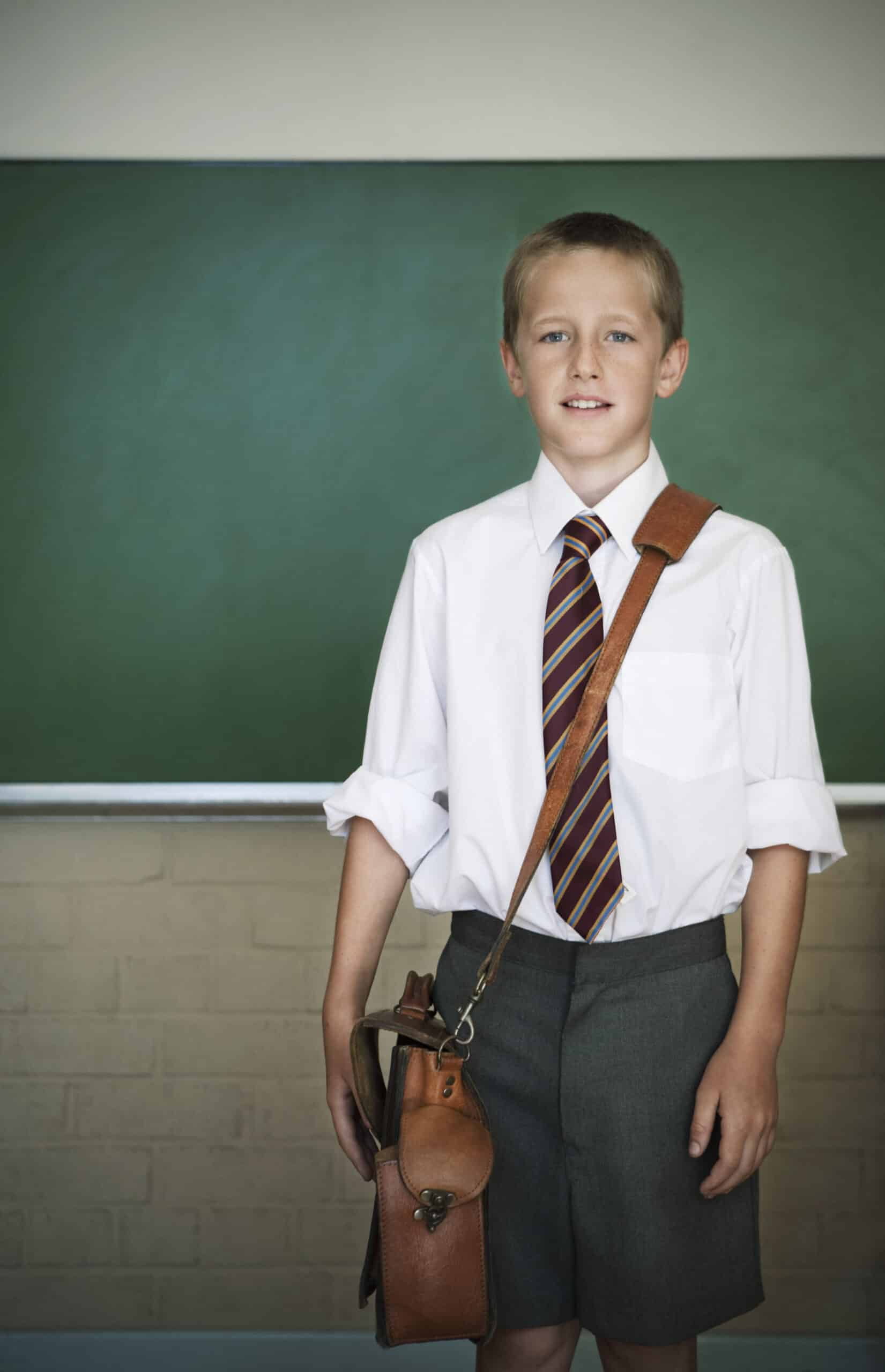 Ein kleiner Junge steht vor einer grünen Tafel und ist bereit für die Schule. Er trägt ein weißes Oberhemd, eine gestreifte Krawatte, graue Shorts und hat eine Ledertasche über der Schulter. Seine Ärmel sind hochgekrempelt und er lächelt leicht und blickt direkt in die Kamera. © Fotografie Tomas Rodriguez