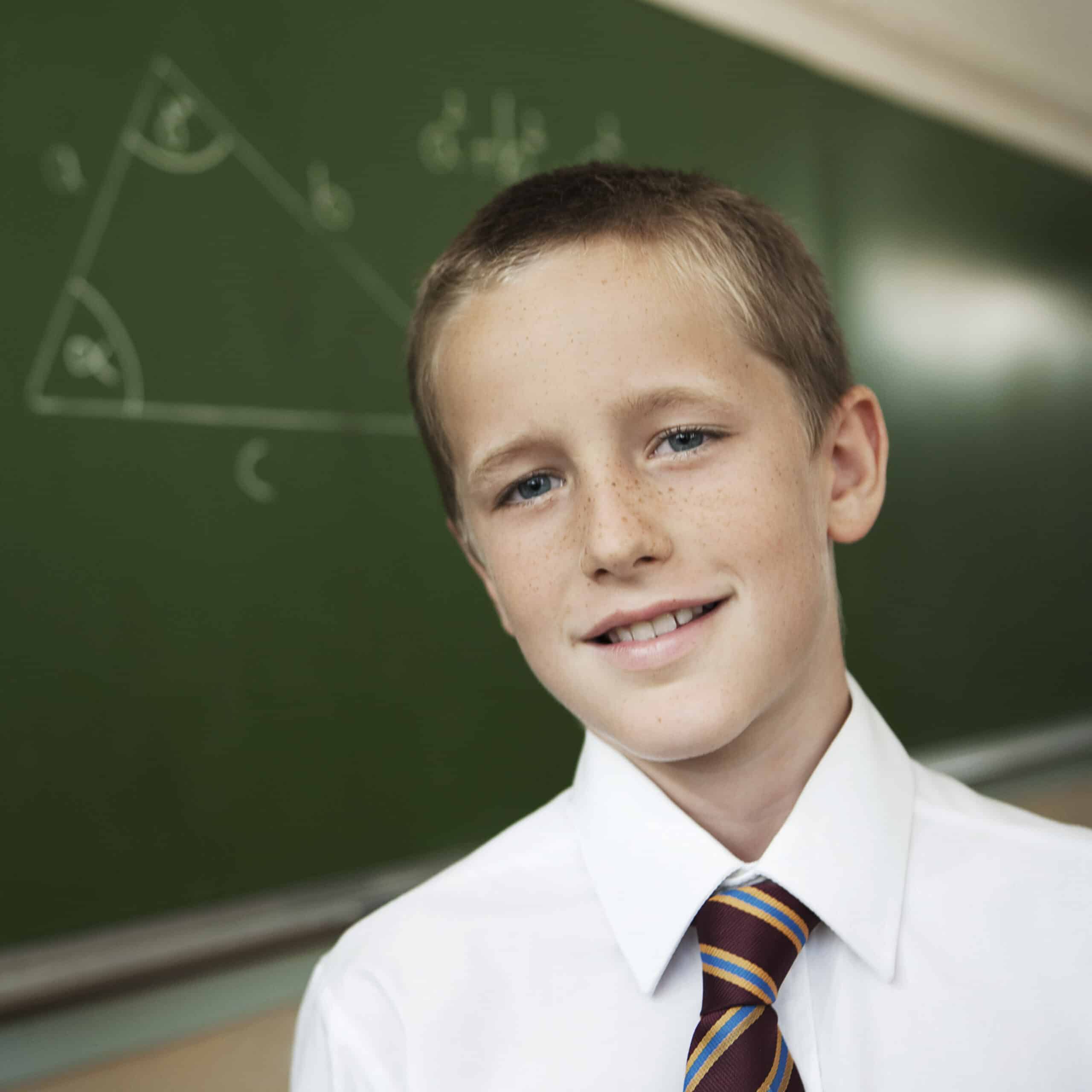 Ein kleiner Junge mit kurzen blonden Haaren, einem weißen Hemd und einer gestreiften Krawatte steht vor einer grünen Tafel, auf der ein Dreieck und mathematische Gleichungen gezeichnet sind. Er lächelt leicht und blickt in die Kamera. © Fotografie Tomas Rodriguez