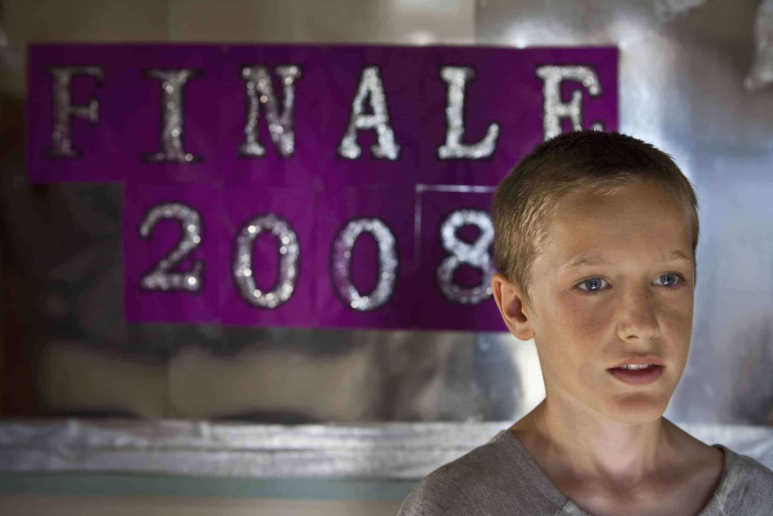 Ein kleiner Junge mit kurzen Haaren steht vor einem lila-silbernen Schild mit der Aufschrift „FINALE 2008“. Er trägt ein graues Hemd und schaut leicht zur Seite. Der Hintergrund scheint reflektierend und bringt etwas Licht in das Bild. © Fotografie Tomas Rodriguez