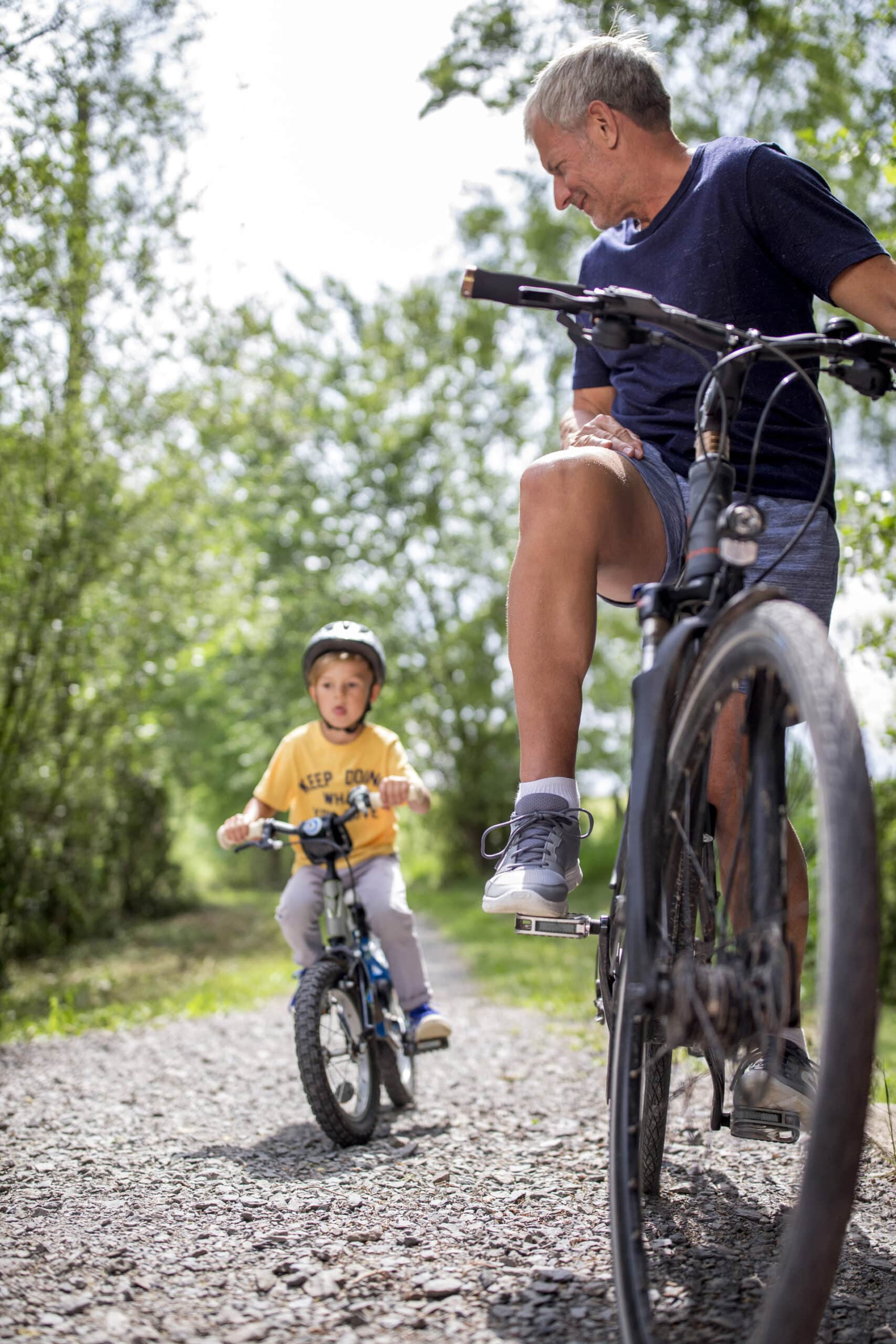 Ein älterer Erwachsener und ein kleines Kind fahren auf einem von Bäumen umgebenen Schotterweg Fahrrad. Das Kind, das einen Helm trägt, sitzt auf einem kleineren Fahrrad und blickt zu dem Erwachsenen auf, der lächelt und auf einem größeren Fahrrad balanciert. Die Szene ist sonnig und hell. © Fotografie Tomas Rodriguez
