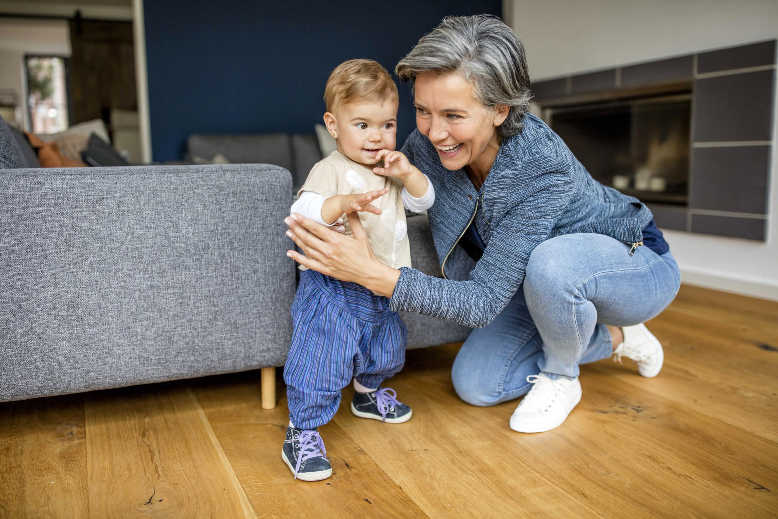 Eine lächelnde Frau mit kurzen grauen Haaren und einem blauen Outfit kniet auf einem Holzboden und führt ein lächelndes Kleinkind, das neben einem grauen Sofa steht. Das Kind trägt ein helles Hemd, blaue Hosen und Turnschuhe. Im Hintergrund sind ein Kamin und einige Möbel zu sehen. © Fotografie Tomas Rodriguez