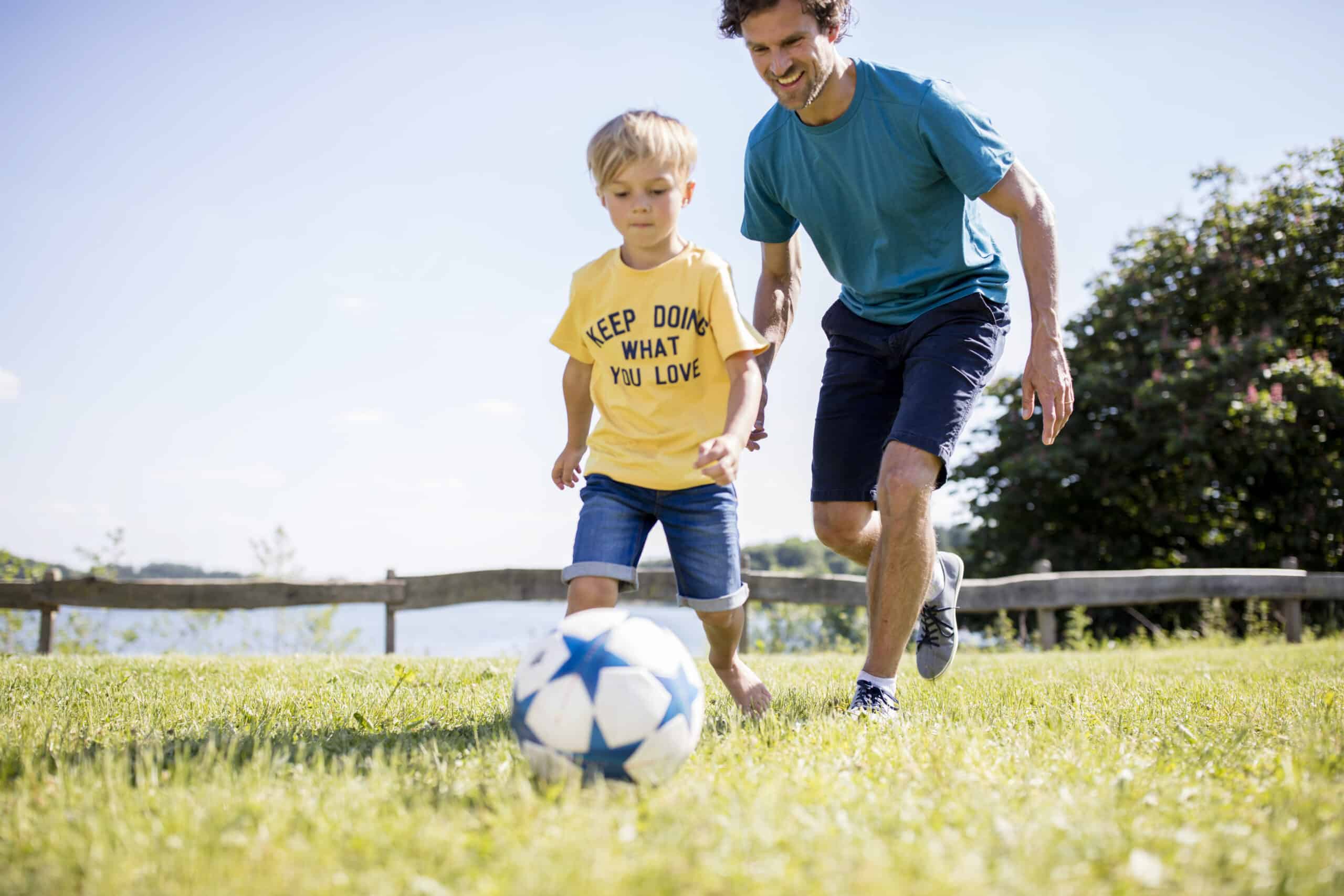 Ein Mann und ein kleiner Junge spielen auf einem sonnigen Rasenplatz Fußball. Der Junge, der ein gelbes T-Shirt mit der Aufschrift „MACH WEITER, WAS DU LIEBST“ trägt, rennt dem Ball hinterher. Der Mann, der ein blaues T-Shirt und Shorts trägt, lächelt und folgt ihm dicht auf den Fersen. © Fotografie Tomas Rodriguez