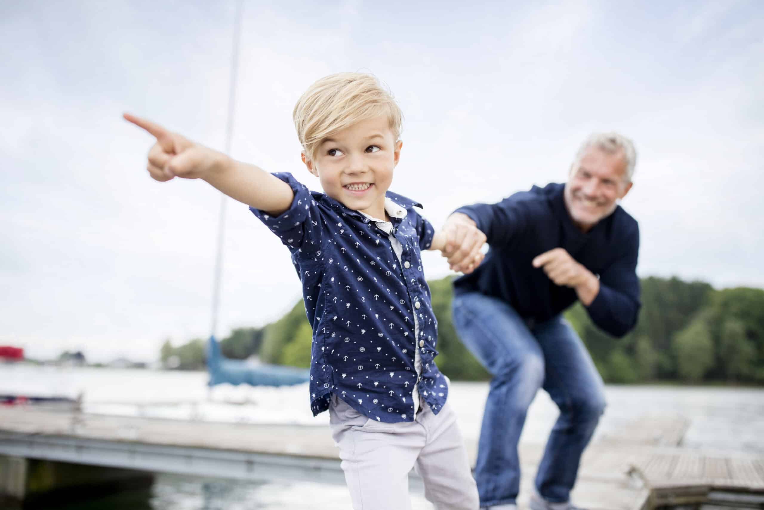 Ein kleiner Junge mit blondem Haar, der ein marineblaues Hemd mit weißen Punkten trägt, zeigt aufgeregt in die Ferne, während er die Hand eines älteren Mannes mit grauem Haar hält. Sie stehen auf einem Steg mit Wasser und Bäumen im Hintergrund. © Fotografie Tomas Rodriguez