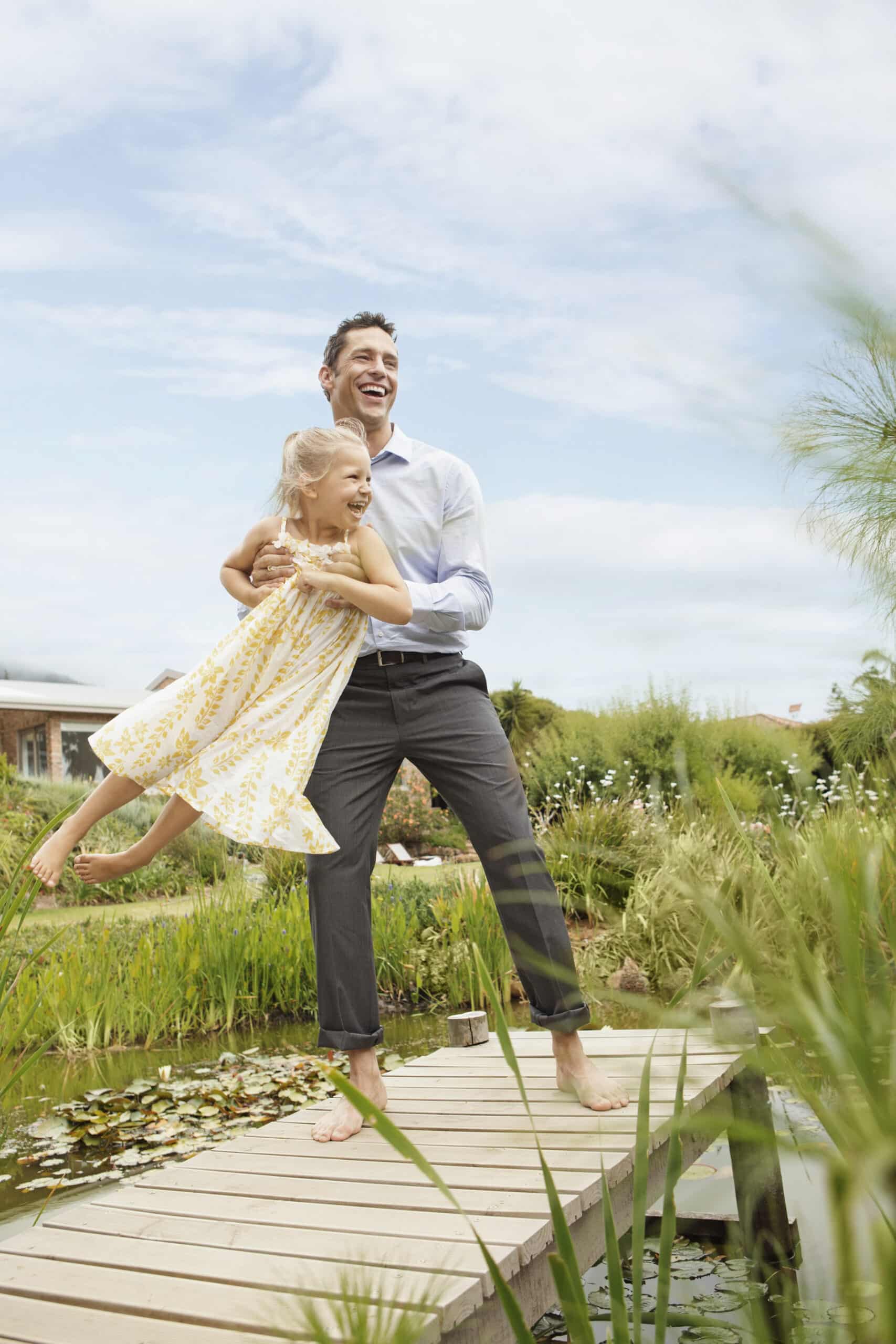 Ein Mann in weißem Hemd und grauer Hose hält fröhlich ein junges Mädchen in einem gelben Kleid, das zu lachen scheint. Sie stehen barfuß auf einer Holzbrücke über einem Teich mit Seerosenblättern, im Hintergrund sind Grünpflanzen und ein Haus unter einem blauen Himmel zu sehen. © Fotografie Tomas Rodriguez