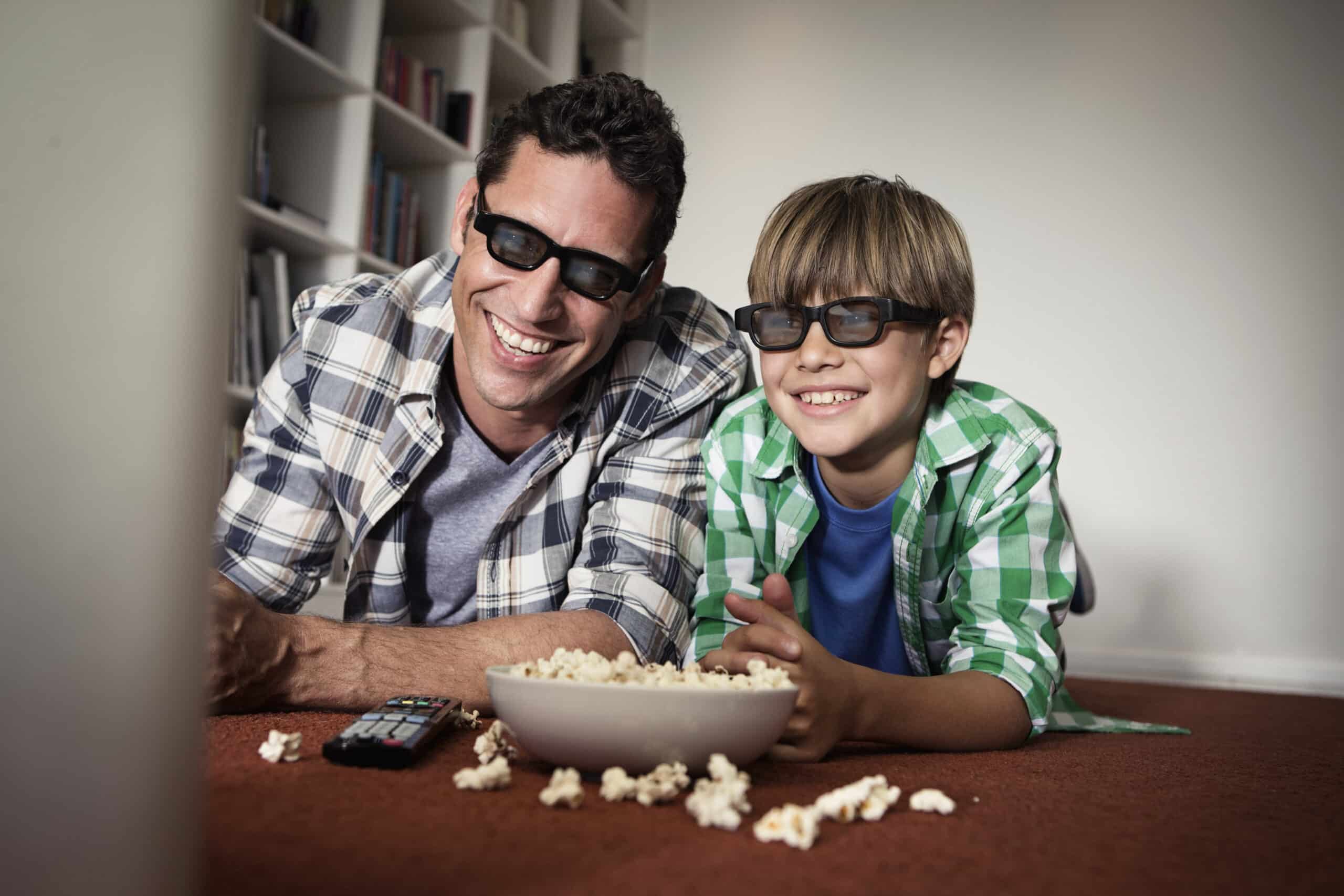 Ein Mann und ein kleiner Junge mit 3D-Brille liegen lächelnd auf dem Teppich und schauen sich einen Film an. Zwischen ihnen steht eine Schüssel Popcorn und sie befinden sich in einem gemütlichen Raum mit einem Bücherregal im Hintergrund. © Fotografie Tomas Rodriguez