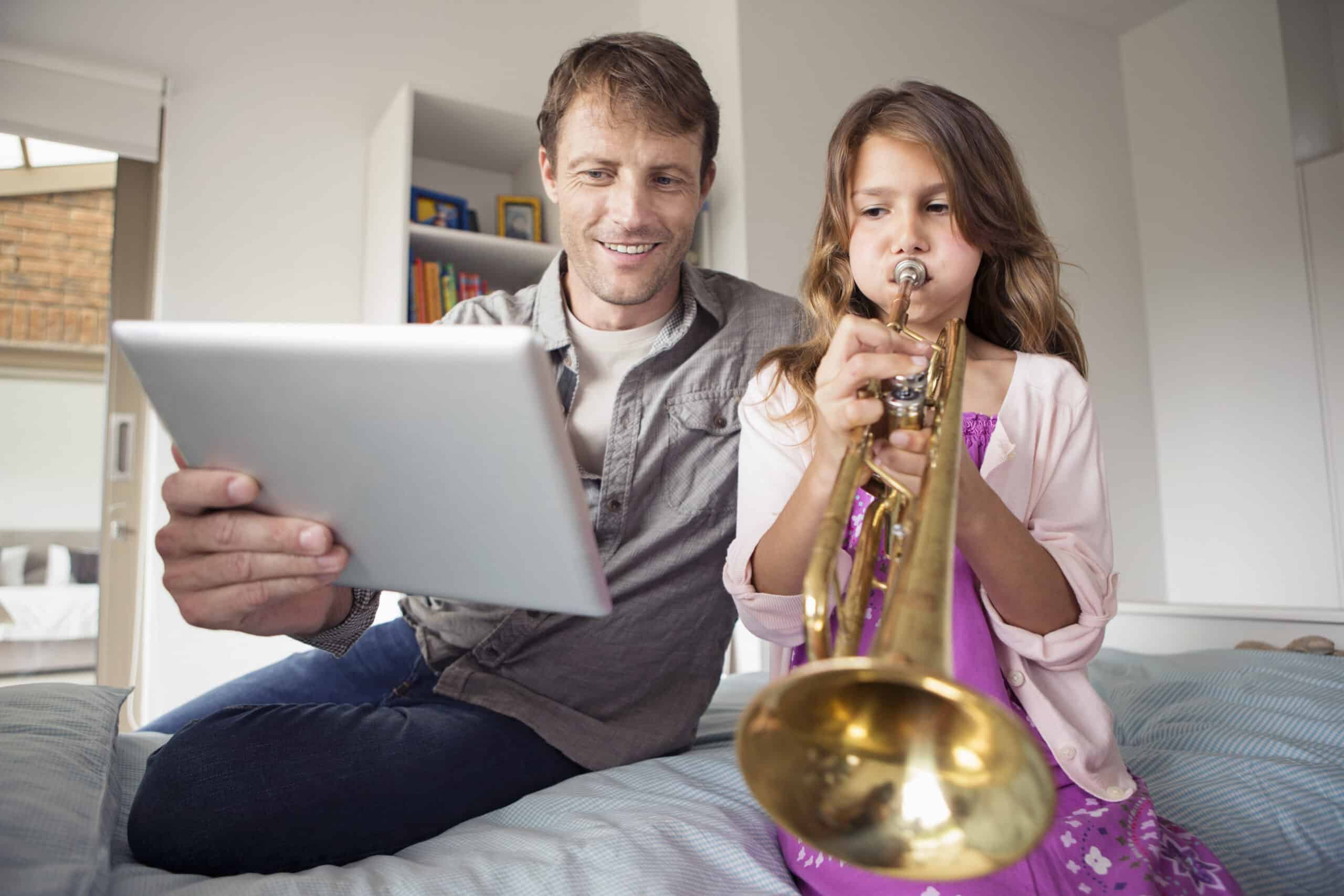 Ein Mann und ein junges Mädchen sitzen auf einem Bett. Der Mann hält ein Tablet und lächelt. Das Mädchen spielt Trompete und konzentriert sich auf das Instrument. Sie befinden sich in einem hellen, gemütlichen Raum mit Bücherregalen und einem Fenster im Hintergrund. © Fotografie Tomas Rodriguez
