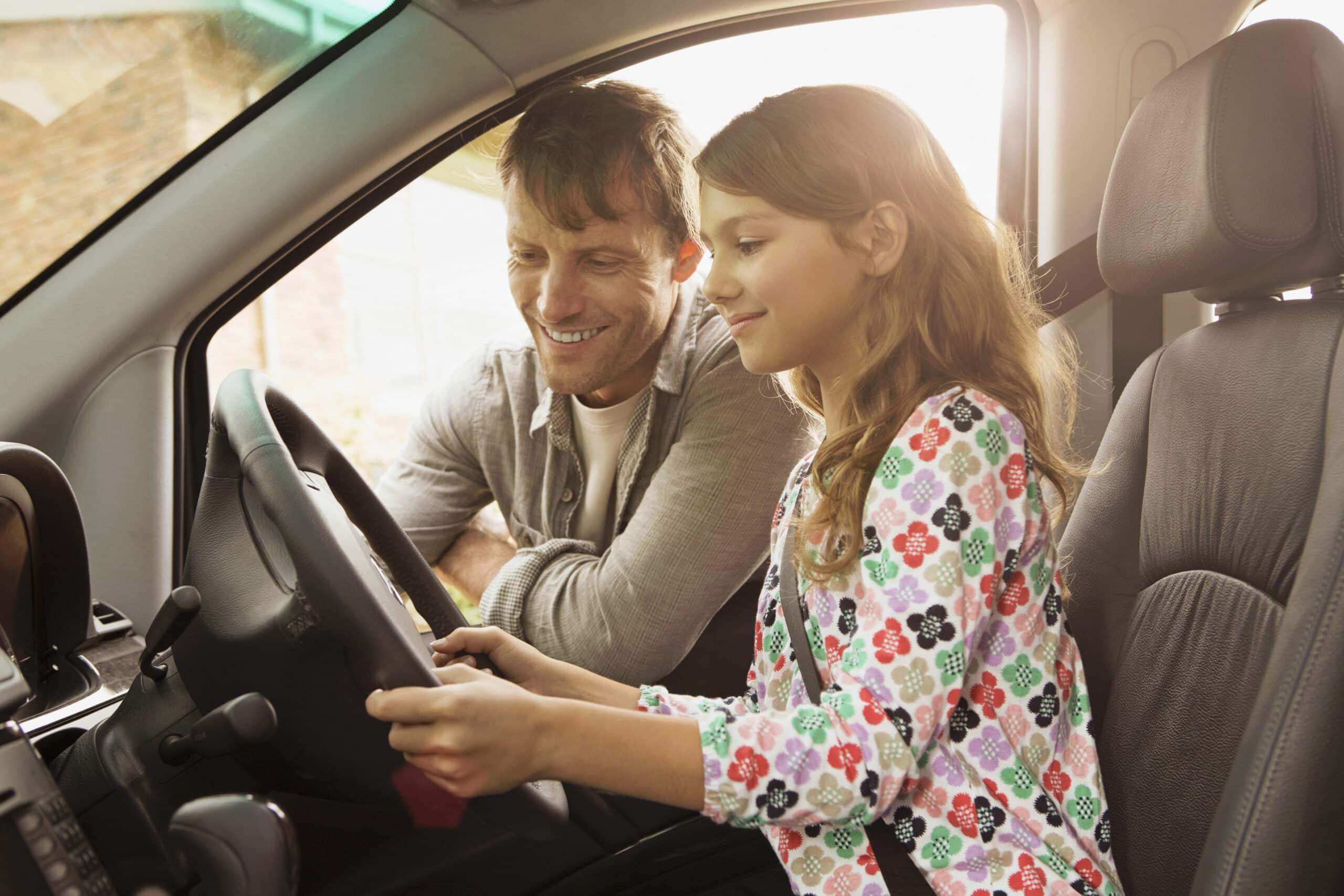 Ein Mann und ein junges Mädchen sitzen in einem Auto. Der Mann auf dem Beifahrersitz lächelt und schaut das Mädchen an, das auf dem Fahrersitz sitzt und das Lenkrad mit beiden Händen hält. Sonnenlicht fällt durch die Fenster und schafft eine warme Atmosphäre. © Fotografie Tomas Rodriguez