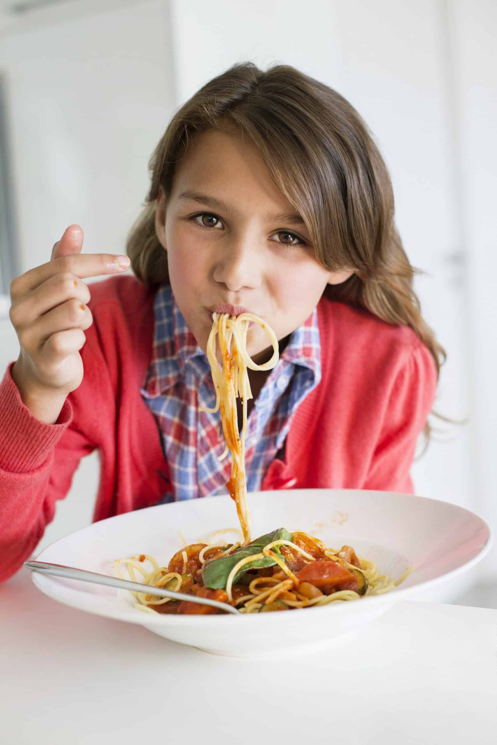 Ein junges Mädchen mit langen braunen Haaren, einer roten Strickjacke und einem karierten Hemd sitzt an einem Tisch und isst Spaghetti von einem weißen Teller. Sie hält die Spaghetti mit ihrer Gabel und führt sie zum Mund, wobei ein Stück Spaghetti heraushängt. © Fotografie Tomas Rodriguez