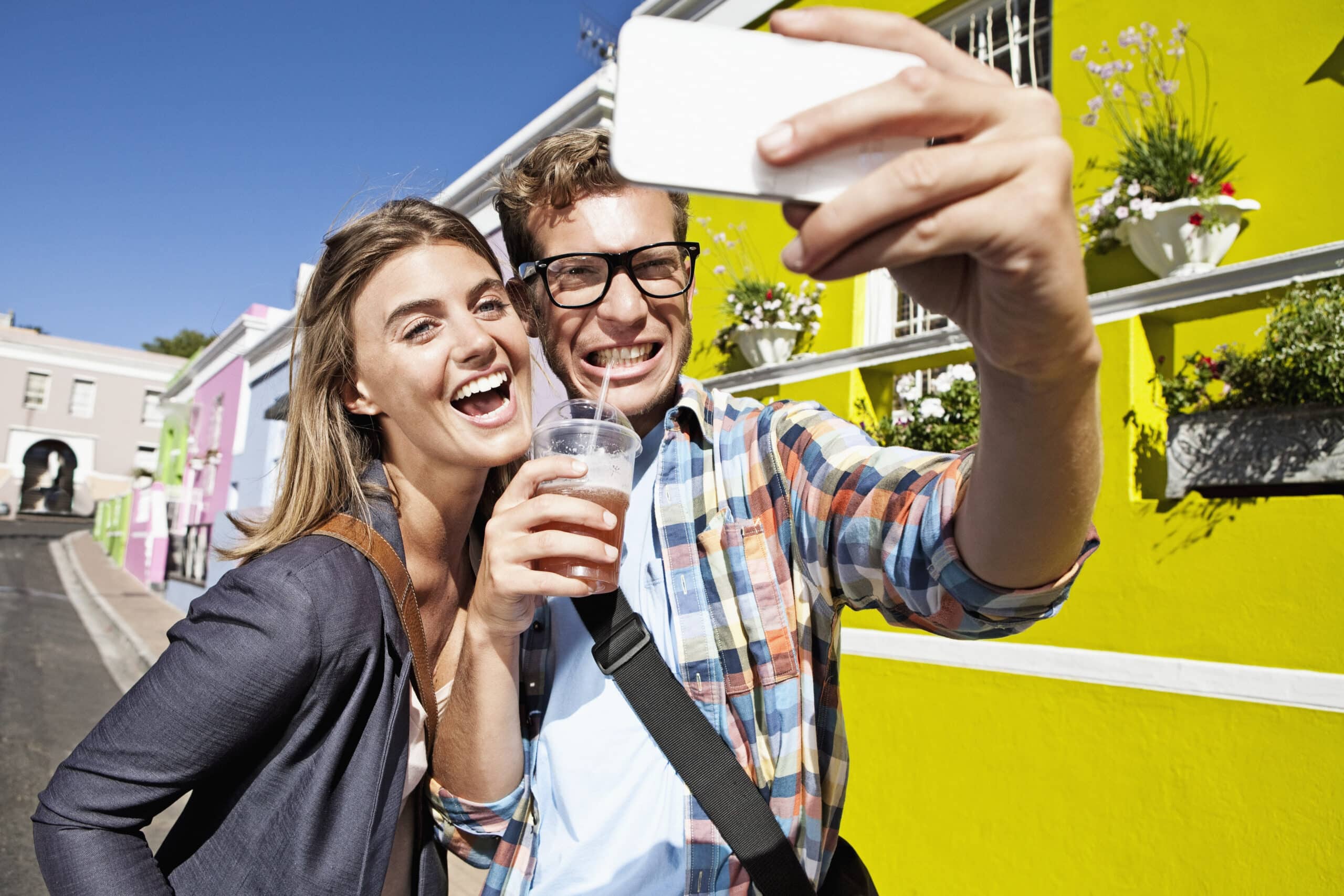 Ein fröhliches junges Paar macht auf einer sonnigen Straße mit bunten Gebäuden im Hintergrund ein Selfie mit einem Smartphone. Die Frau hält ein Getränk in der Hand und lehnt sich nah an den Mann heran, der eine Brille trägt. © Fotografie Tomas Rodriguez