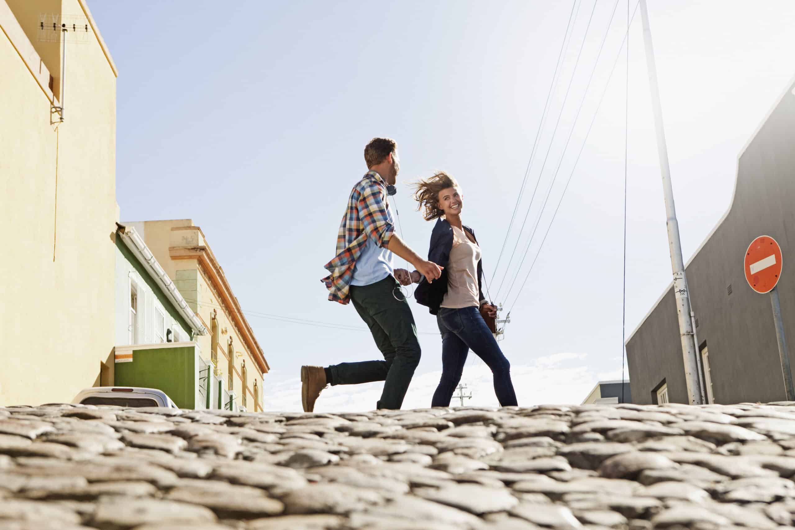 Ein fröhliches junges Paar hält Händchen, während es in einer sonnigen städtischen Umgebung über eine Kopfsteinpflasterstraße geht. Im Hintergrund sind Gebäude und ein Stoppschild zu sehen. © Fotografie Tomas Rodriguez