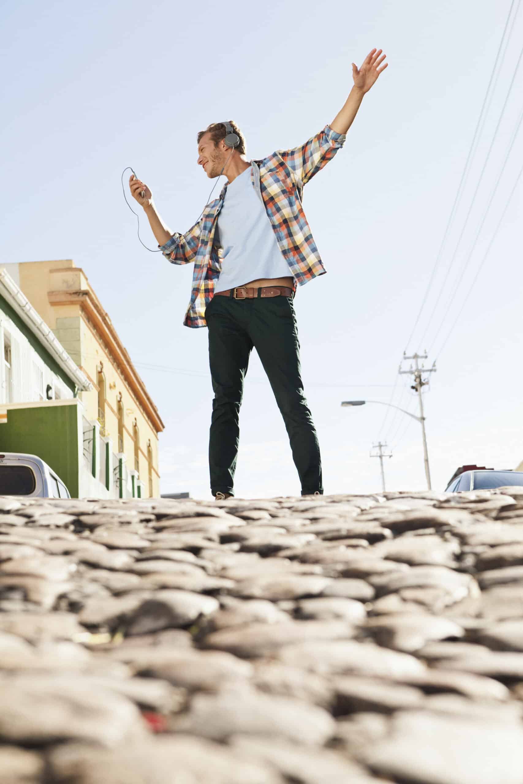 Ein Mann mit Kopfhörern tanzt fröhlich auf einer Kopfsteinpflasterstraße und schwenkt einen Arm in der Luft, über ihm ein klarer blauer Himmel und im Hintergrund Gebäude. © Fotografie Tomas Rodriguez