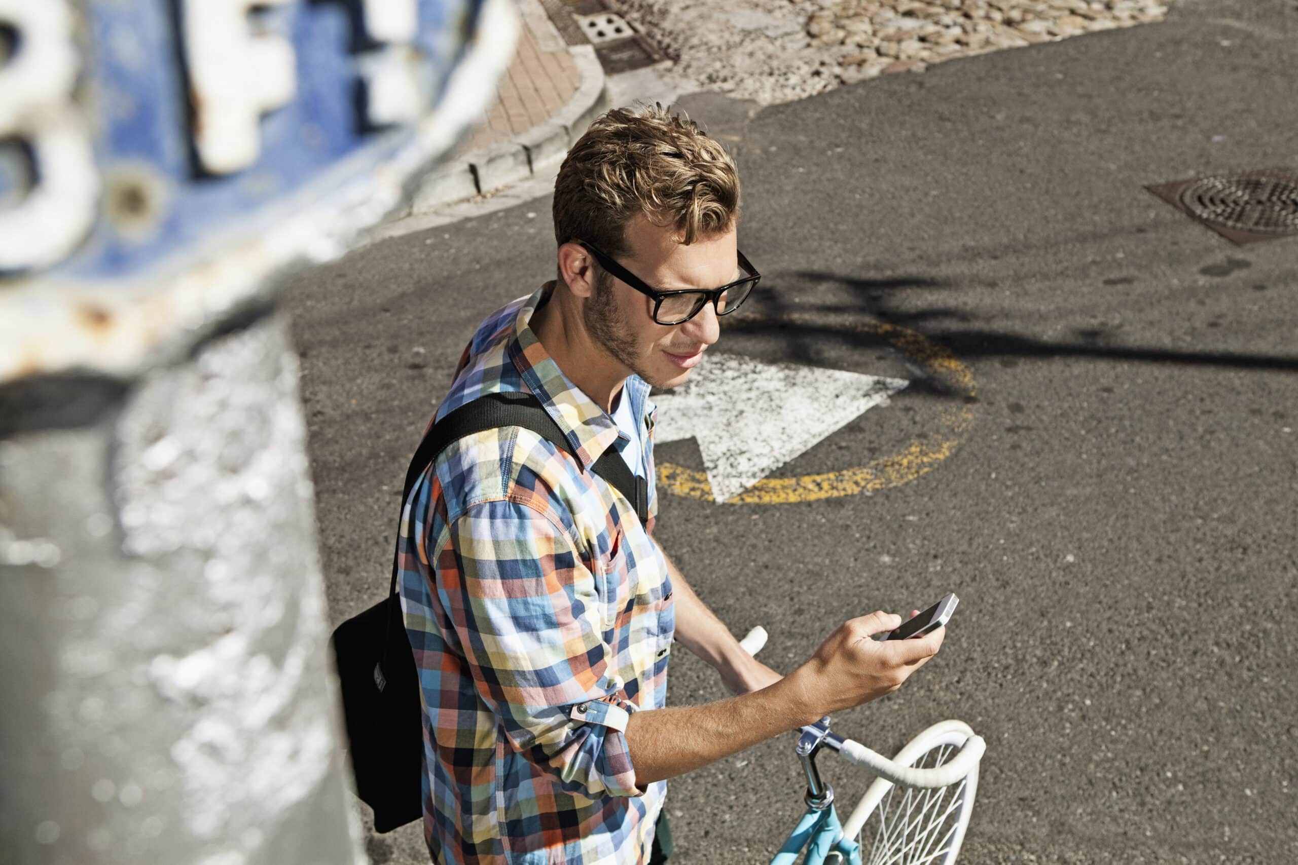 Ein junger Mann mit blonden Haaren, Brille und kariertem Hemd steht auf einer sonnenbeschienenen Straße, schaut auf sein Smartphone, neben ihm steht ein Fahrrad. © Fotografie Tomas Rodriguez