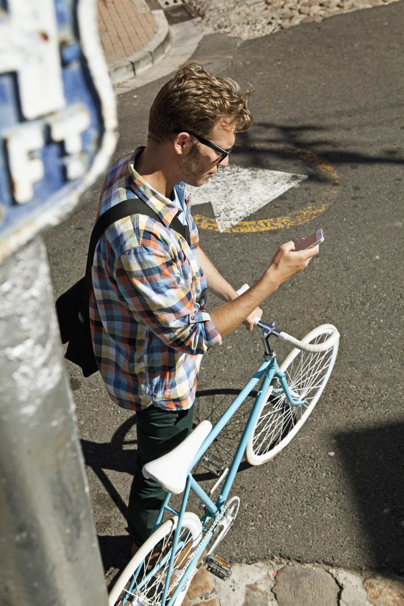 Junger Mann mit Brille und kariertem Hemd hält ein Smartphone und steht neben einem hellblauen Fahrrad auf einer sonnigen Stadtstraße. © Fotografie Tomas Rodriguez