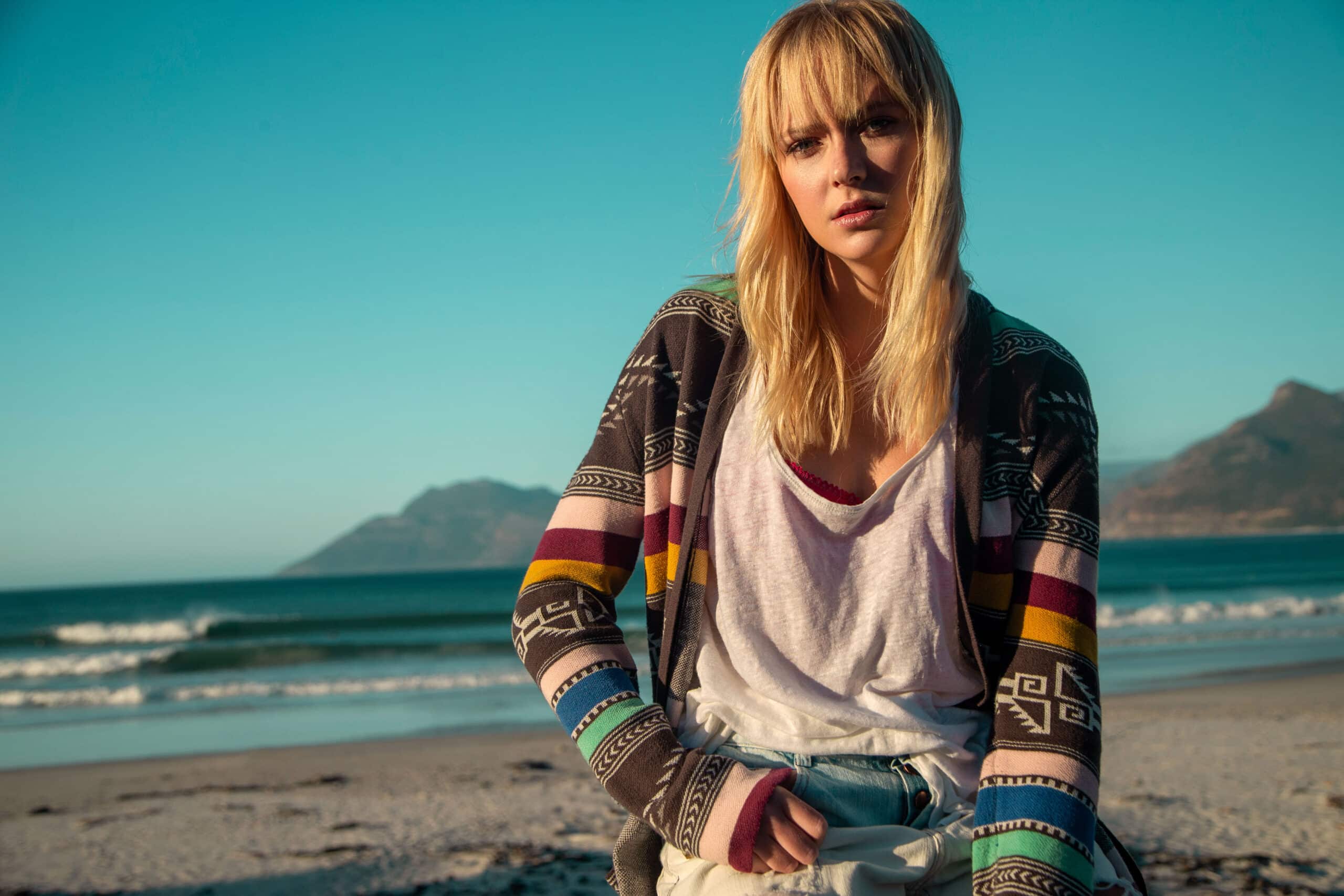 Eine Frau mit blondem Haar, einer Strickjacke mit Tribal-Muster und einem weißen T-Shirt, sitzt an einem Sandstrand, im Hintergrund sind Berge und ein klarer blauer Himmel zu sehen. © Fotografie Tomas Rodriguez