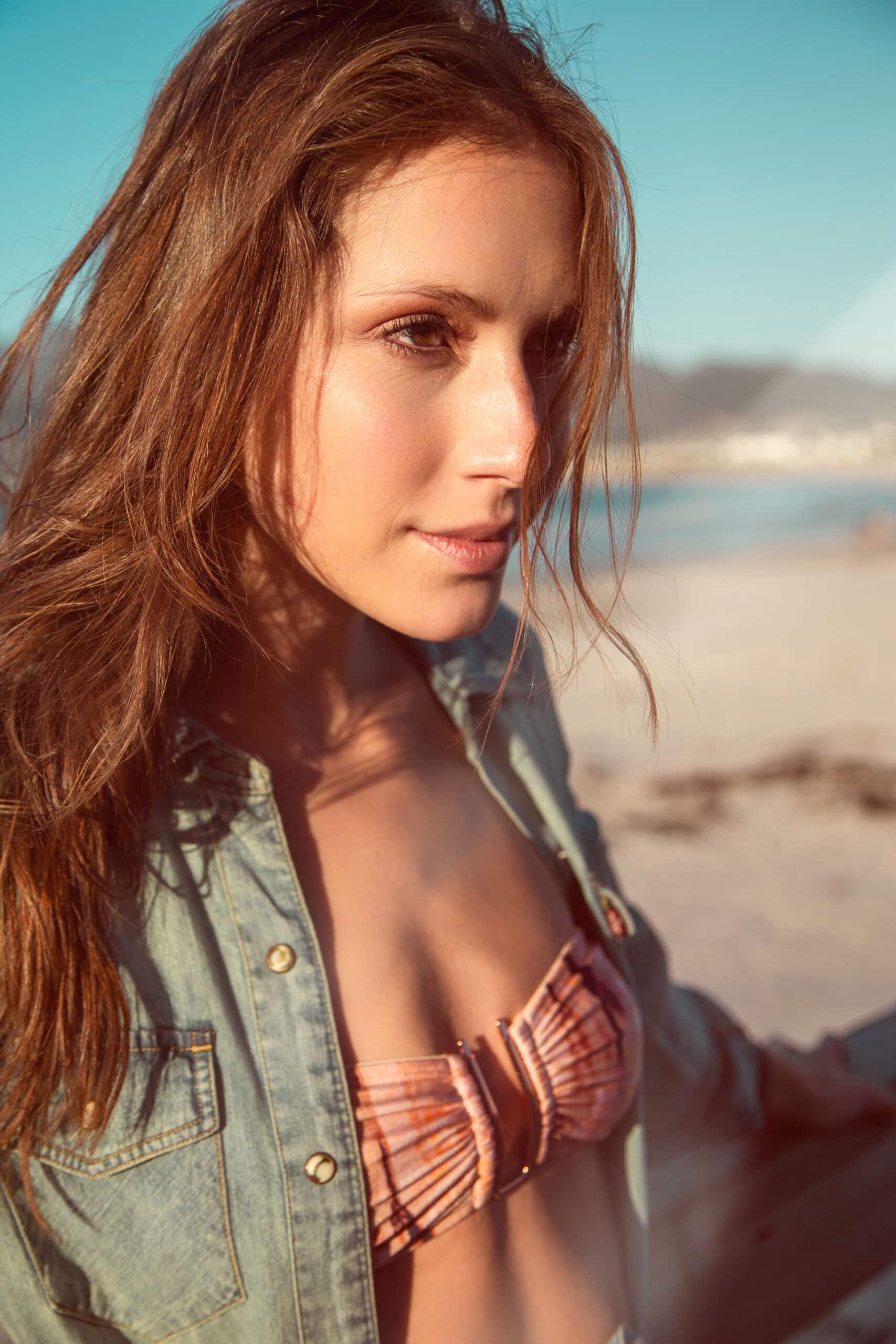 Eine Frau mit langen Haaren, einer Jeansjacke und einem Bikinioberteil trägt, blickt über ihre Schulter, während sie an einem sonnigen Strand steht. Warmes Sonnenlicht betont ihre Gesichtszüge. © Fotografie Tomas Rodriguez