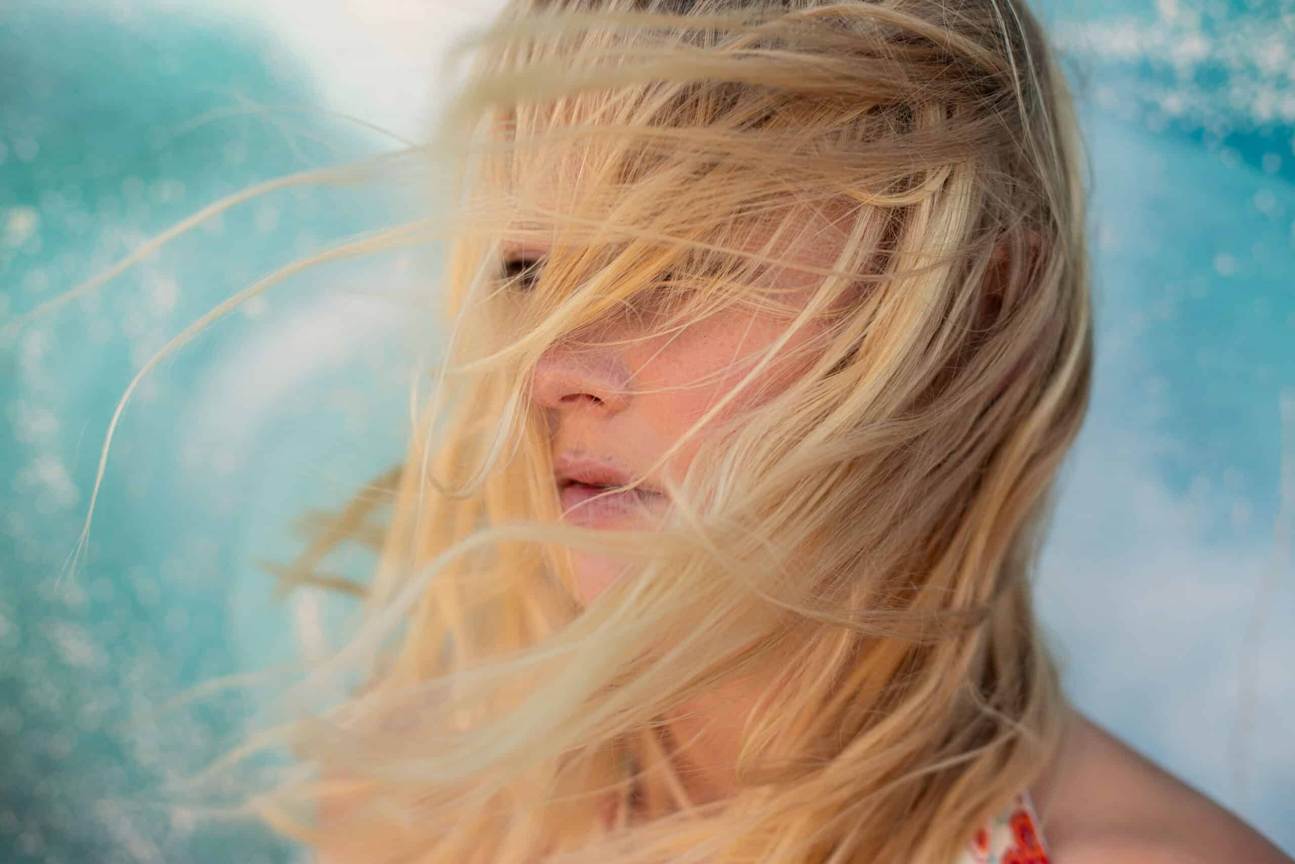Ein Nahaufnahmeporträt einer Person mit langen blonden Haaren, die ihr ins Gesicht wehen, und die vor einem leicht verschwommenen aquamarinfarbenen Hintergrund steht. © Fotografie Tomas Rodriguez