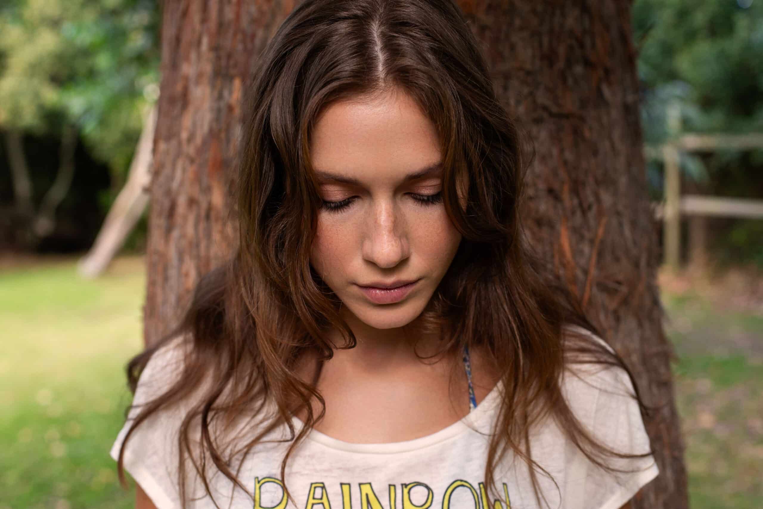 Eine junge Frau mit langen braunen Haaren und einem T-Shirt mit dem Aufdruck „Regenbogen“ steht mit geschlossenen Augen an einem Baumstamm in einem üppigen, grünen Park. © Fotografie Tomas Rodriguez