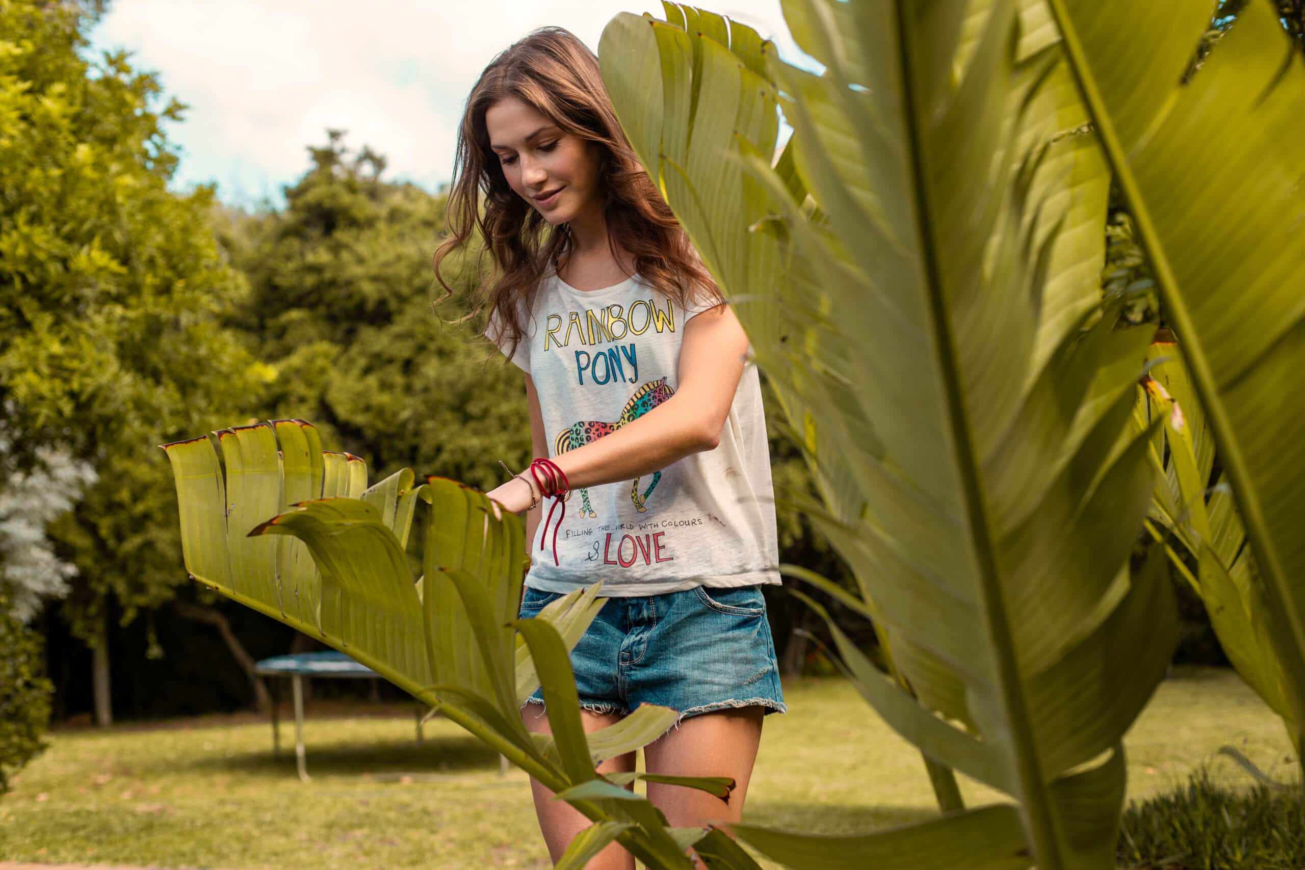 Eine junge Frau in einem lässigen Tanktop und Jeansshorts geht durch einen Garten und berührt sanft ein großes Blatt vor der Kulisse üppigen Grüns. © Fotografie Tomas Rodriguez