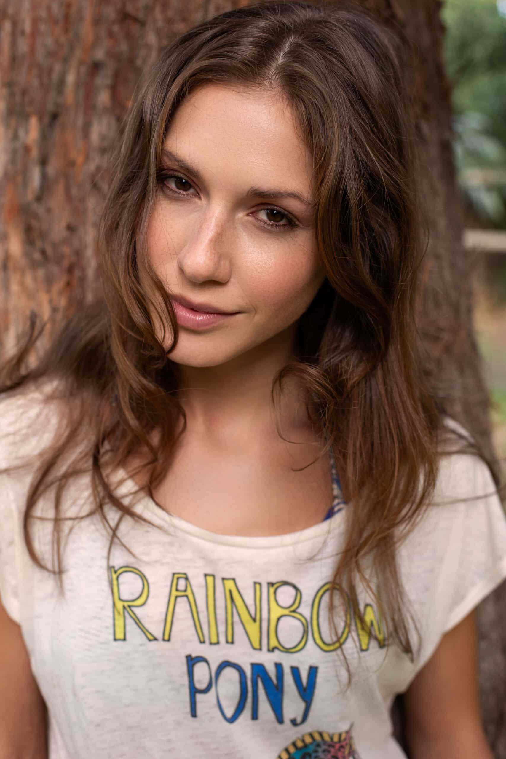 Eine Frau mit braunem Haar und einem subtilen Lächeln lehnt an einem Baum und trägt ein weißes T-Shirt mit der Aufschrift „Regenbogenpony“. © Fotografie Tomas Rodriguez