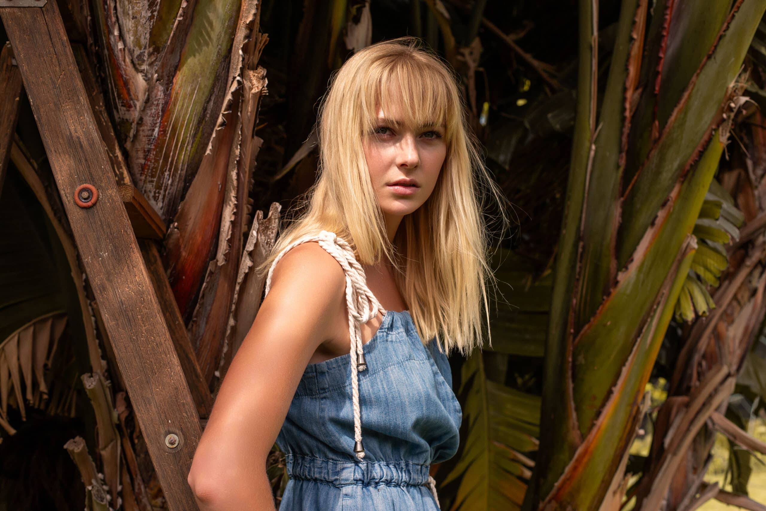 Eine junge Frau mit blonden Haaren und einem Jeanskleid steht zwischen großen tropischen Blättern und blickt aufmerksam in die Kamera. © Fotografie Tomas Rodriguez