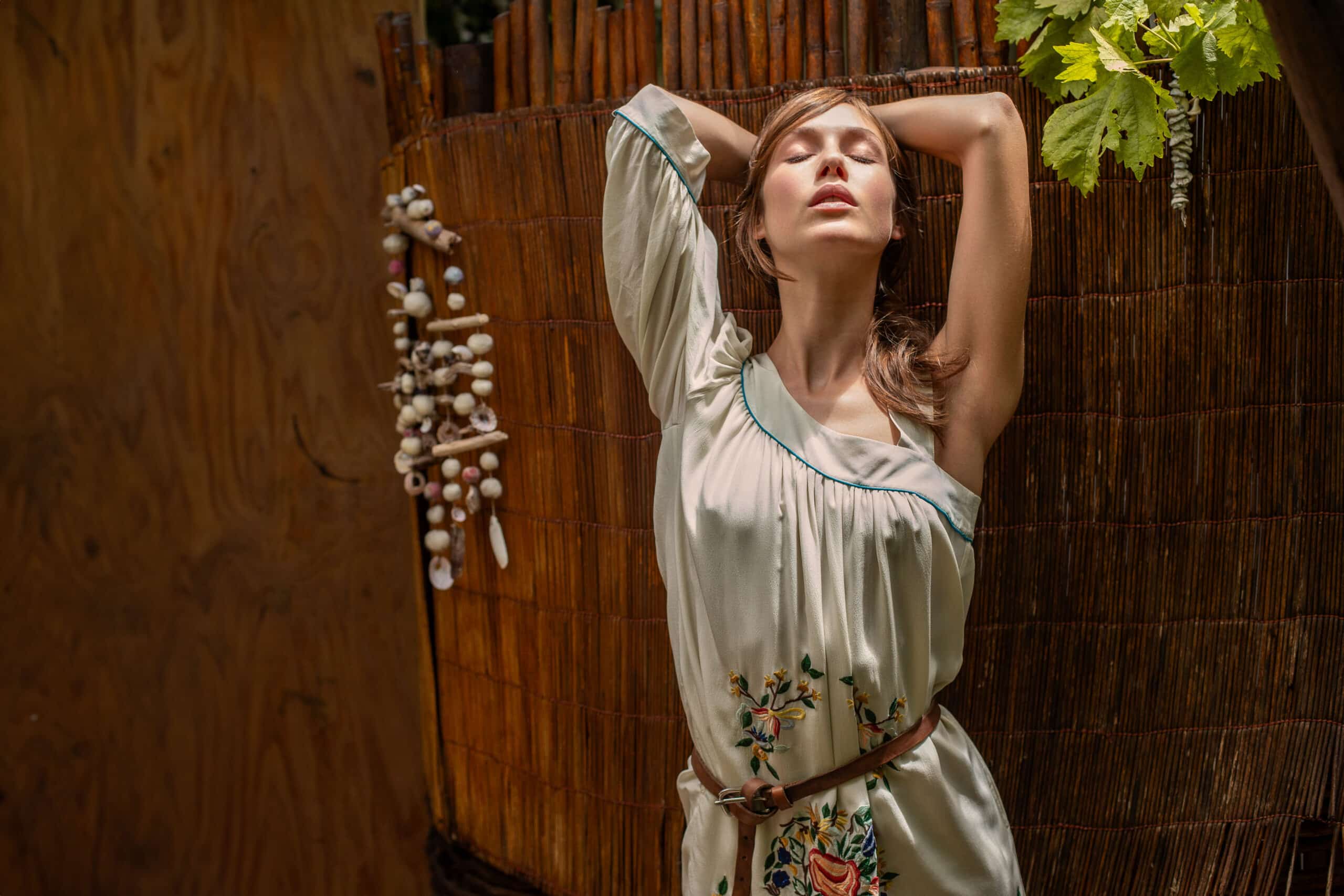 Eine Frau in einem geblümten Kleid steht vor einem Bambushintergrund, schließt die Augen und hebt in einer entspannten Pose die Arme über den Kopf. Natürliches Licht unterstreicht ihren heiteren Gesichtsausdruck. © Fotografie Tomas Rodriguez