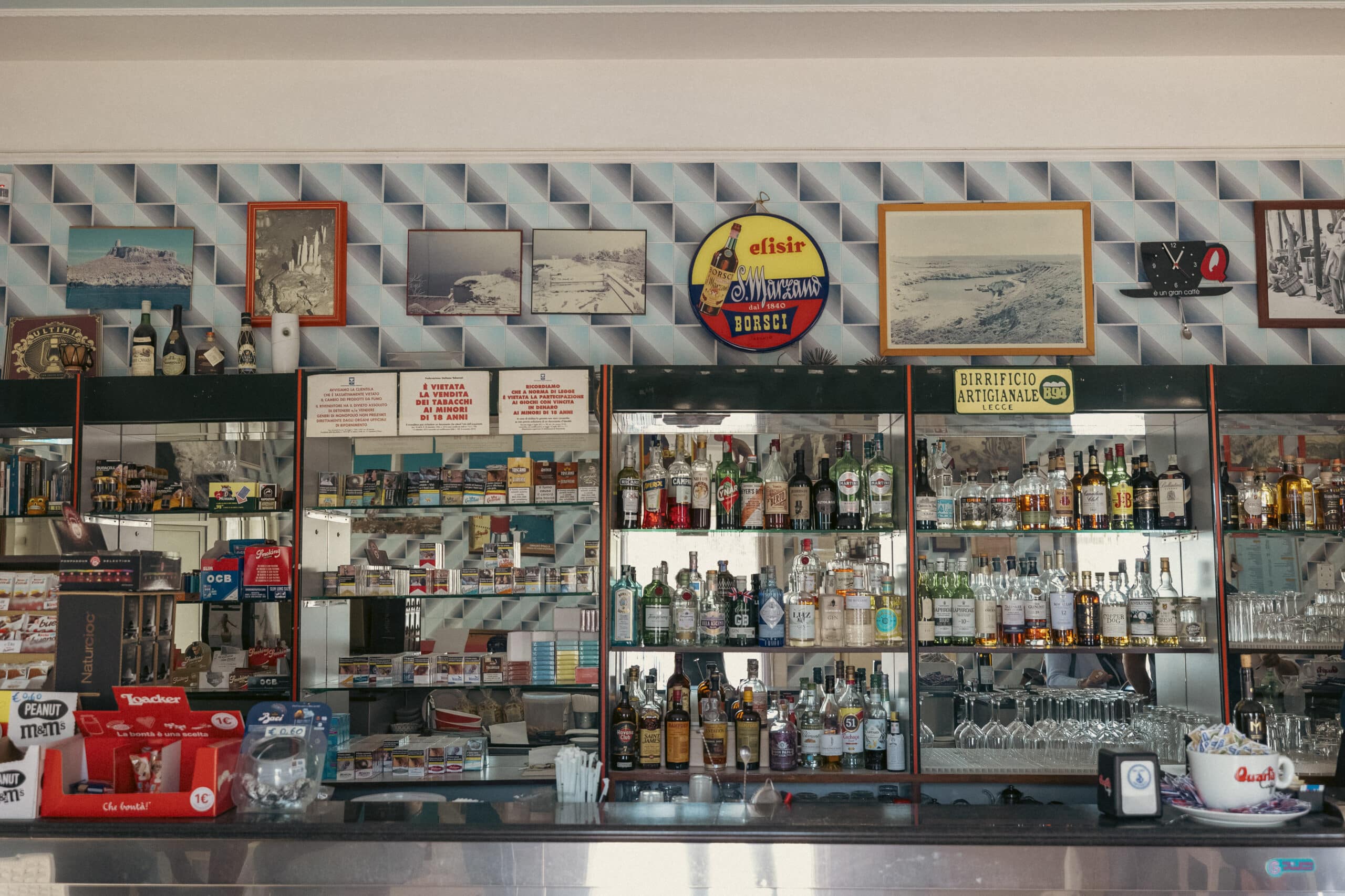 Innenansicht einer Bar im Vintage-Stil mit einer Auswahl an Schnapsflaschen hinter der Theke, Retro-Dekor, gerahmten Bildern an der Wand und einer sichtbaren Menütafel. © Fotografie Tomas Rodriguez