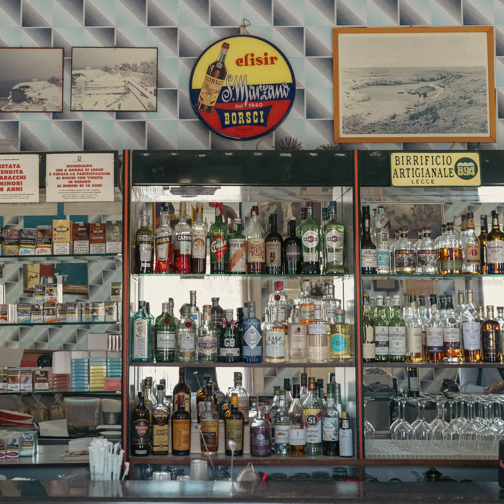 Im Inneren einer Bar werden auf Glasregalen eine Reihe von Schnapsflaschen präsentiert. Zur Einrichtung gehören gerahmte Fotografien, Vintage-Schilder und ein strukturierter Fliesenhintergrund, der ein Retro-Ambiente widerspiegelt. © Fotografie Tomas Rodriguez