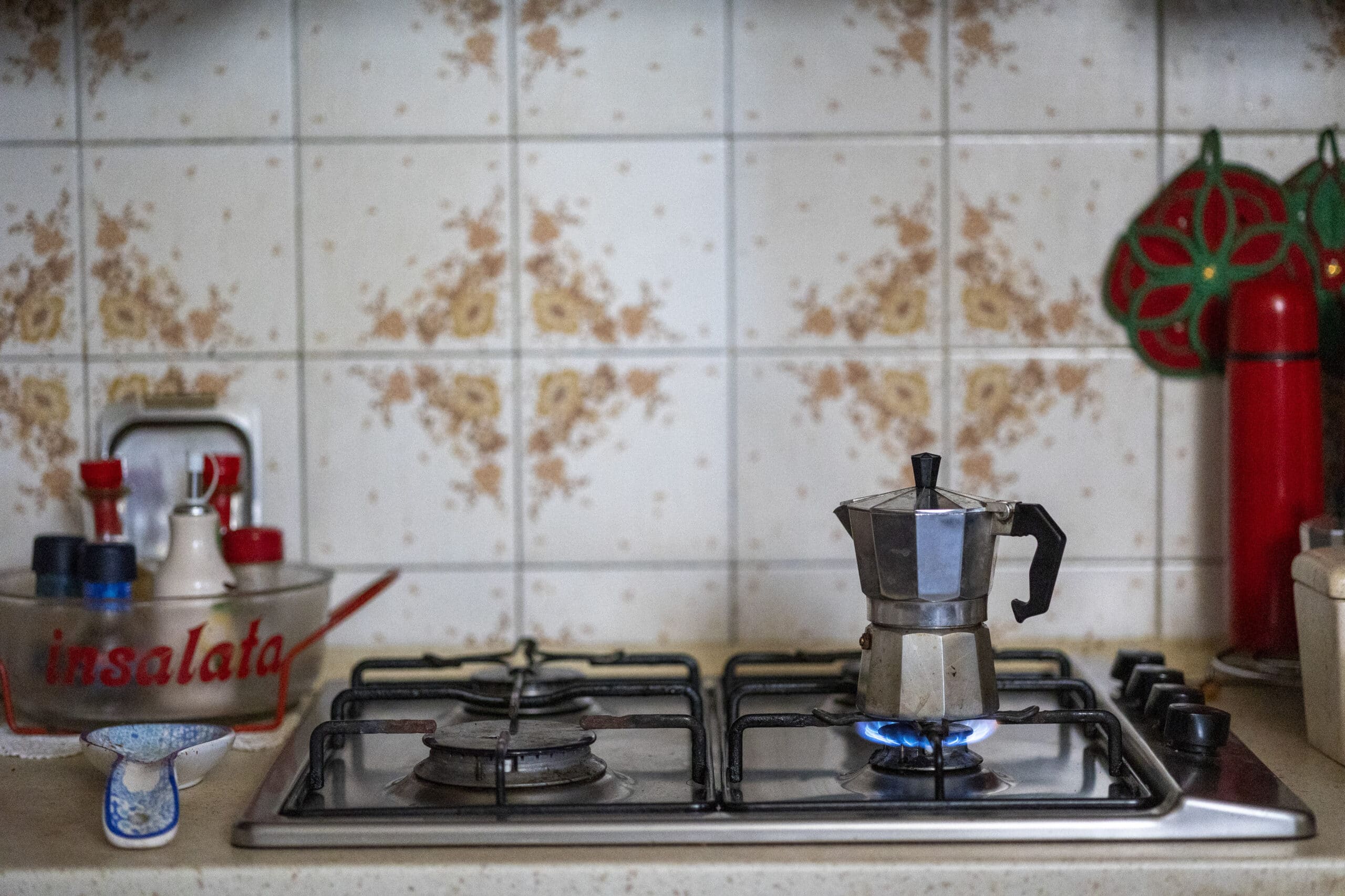 Eine altmodische Herdplatte mit einer metallenen Mokkakanne, die auf einer brennenden Flamme Kaffee kocht, mit Vintage-Küchenutensilien, darunter eine rote „Insalata“-Schüssel, um eine geflieste Küchenrückwand herum. © Fotografie Tomas Rodriguez