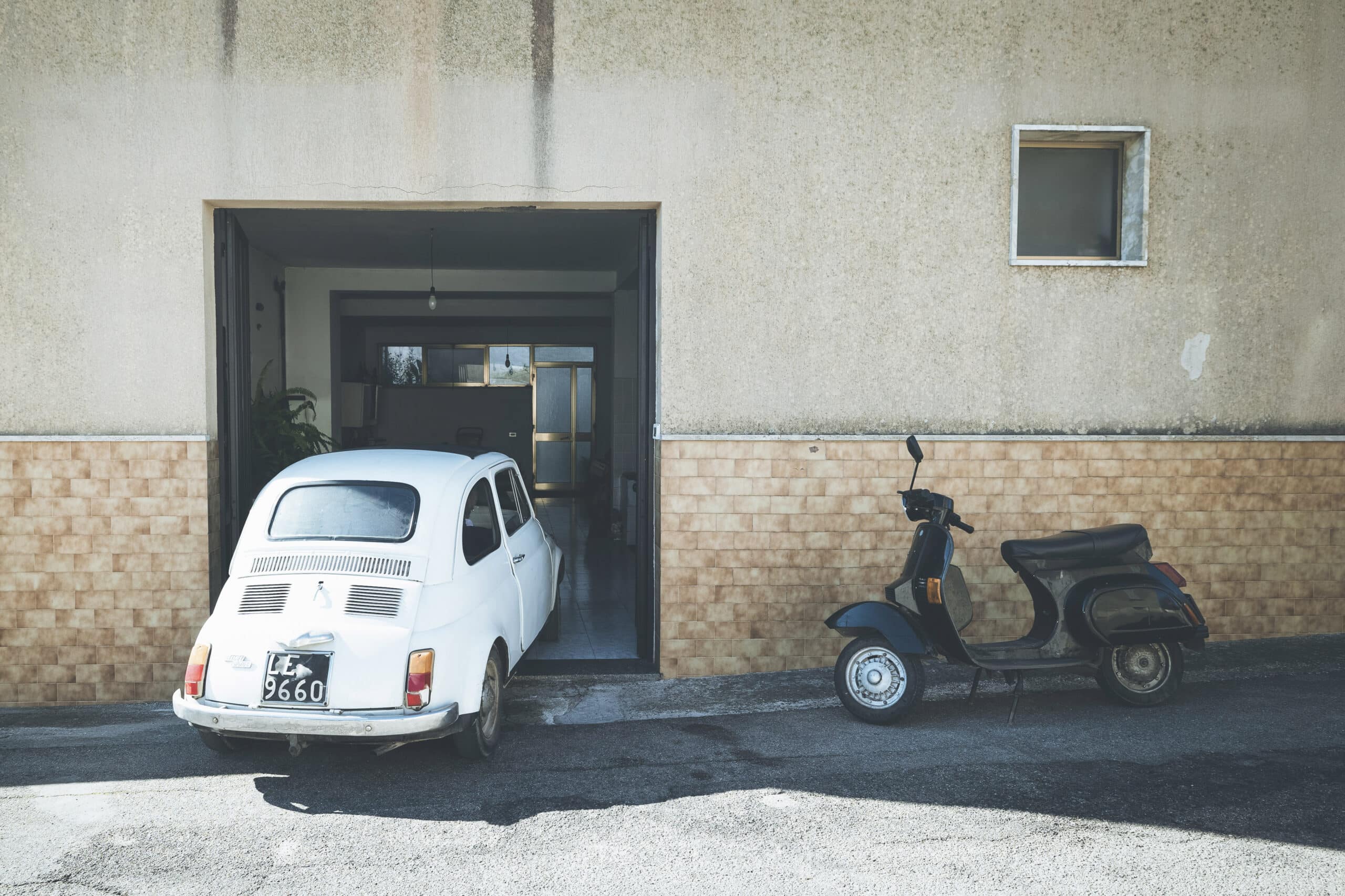 Ein weißer Oldtimer und ein schwarzer Motorroller parken nebeneinander vor einer offenen Garage eines alten Gebäudes mit einer beigen und hellbraunen Fassade. © Fotografie Tomas Rodriguez