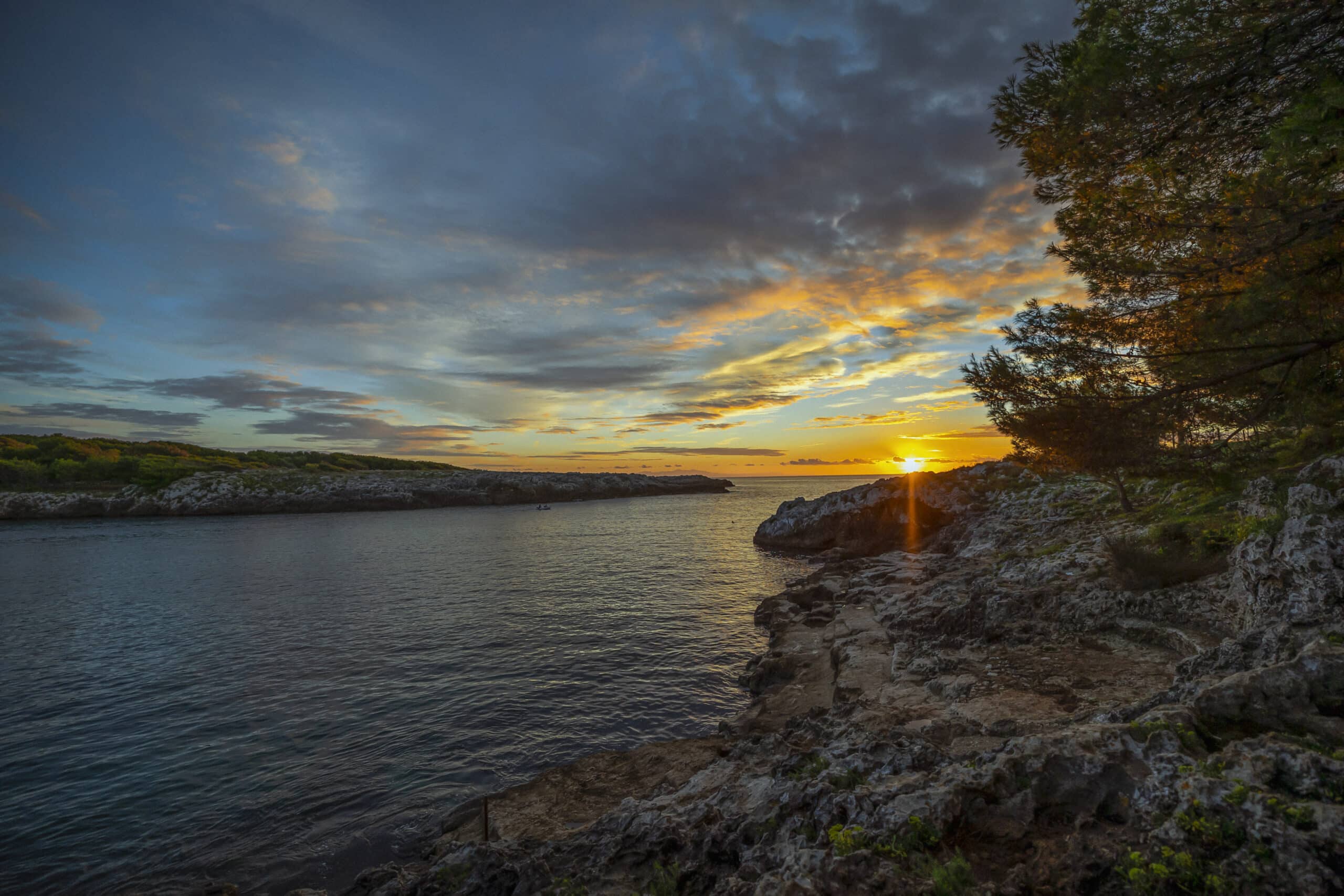 Ein malerischer Sonnenuntergang über einem ruhigen Meer, betrachtet von einer zerklüfteten Küste mit Bäumen. Goldenes Licht erhellt die Wolken und spiegelt sich im Wasser, wodurch die natürliche Schönheit der Landschaft hervorgehoben wird. © Fotografie Tomas Rodriguez