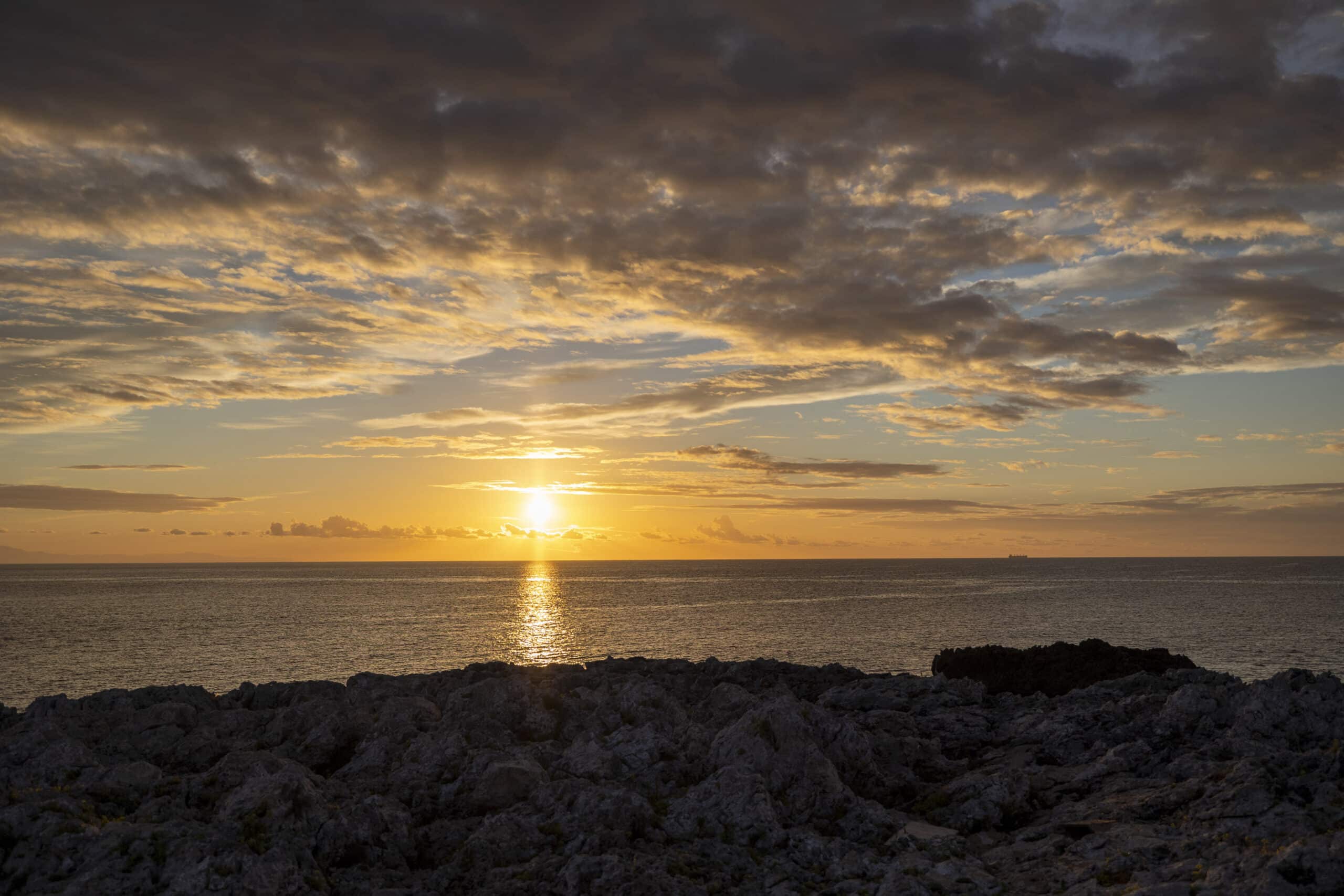 Sonnenuntergang über dem Meer, von einer felsigen Küste aus betrachtet, wobei die Sonne teilweise von Wolken verdeckt wird und ein goldenes Licht auf das Wasser wirft. © Fotografie Tomas Rodriguez