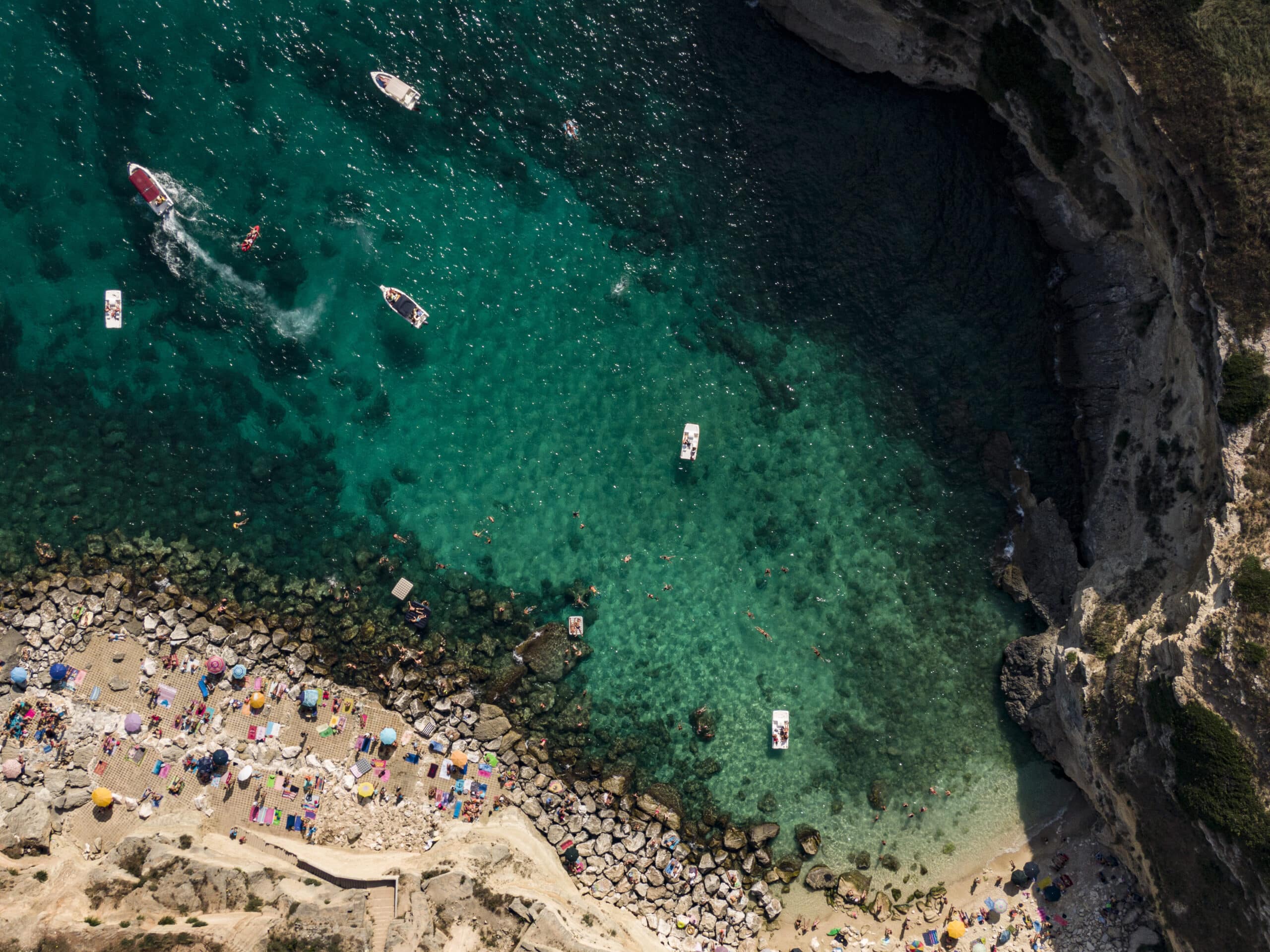 Luftaufnahme eines überfüllten Strandes mit klarem, türkisfarbenem Wasser, umgeben von Klippen, mit schwimmenden und sonnenbadenden Menschen und mehreren kleinen Booten, die in der Nähe schwimmen. © Fotografie Tomas Rodriguez