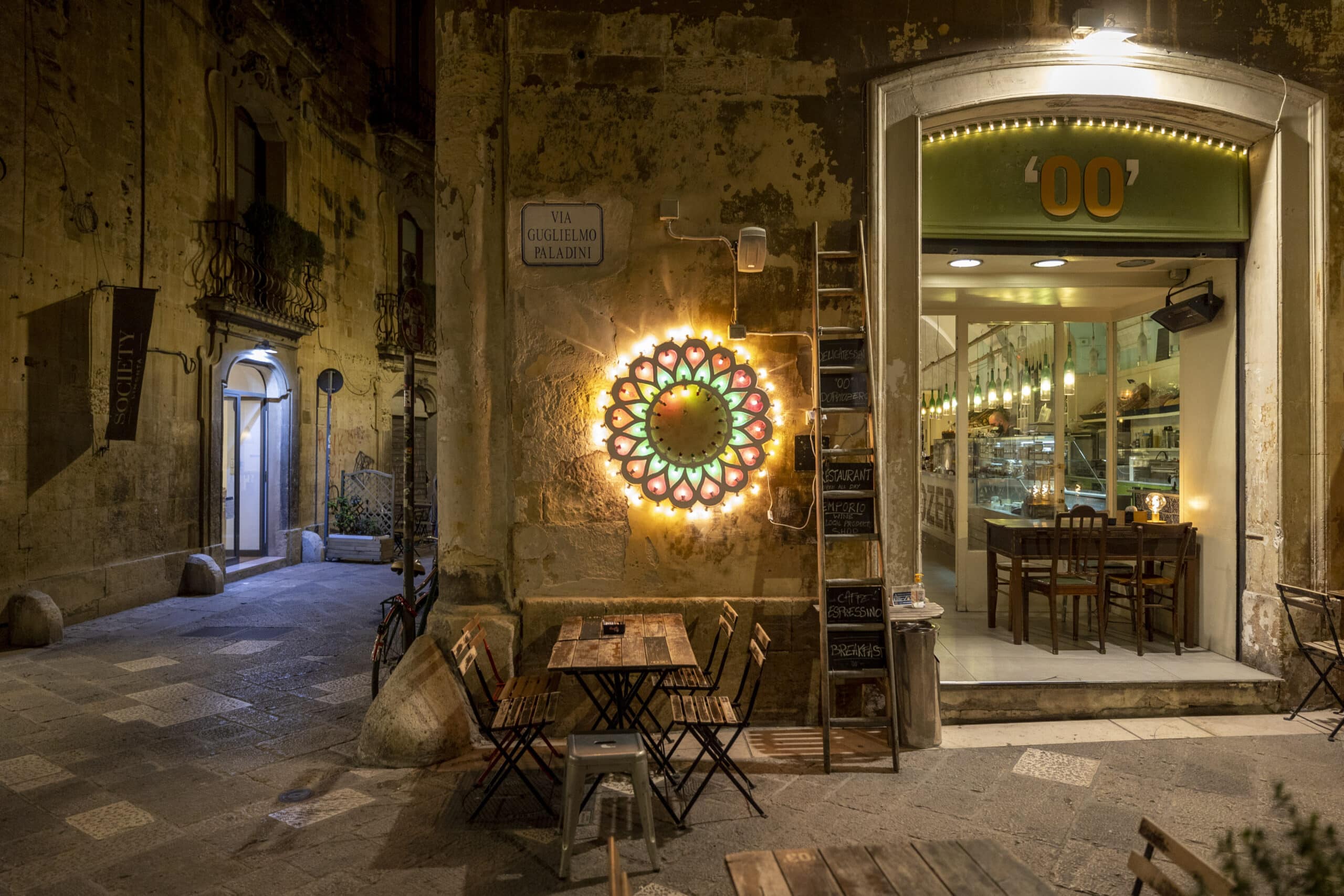 Bezaubernde Straßenansicht bei Nacht mit einem gemütlichen Café mit Sitzgelegenheiten im Freien, einem hellen runden Neonschild und alten strukturierten Gebäudewänden in einer europäischen Gasse. © Fotografie Tomas Rodriguez
