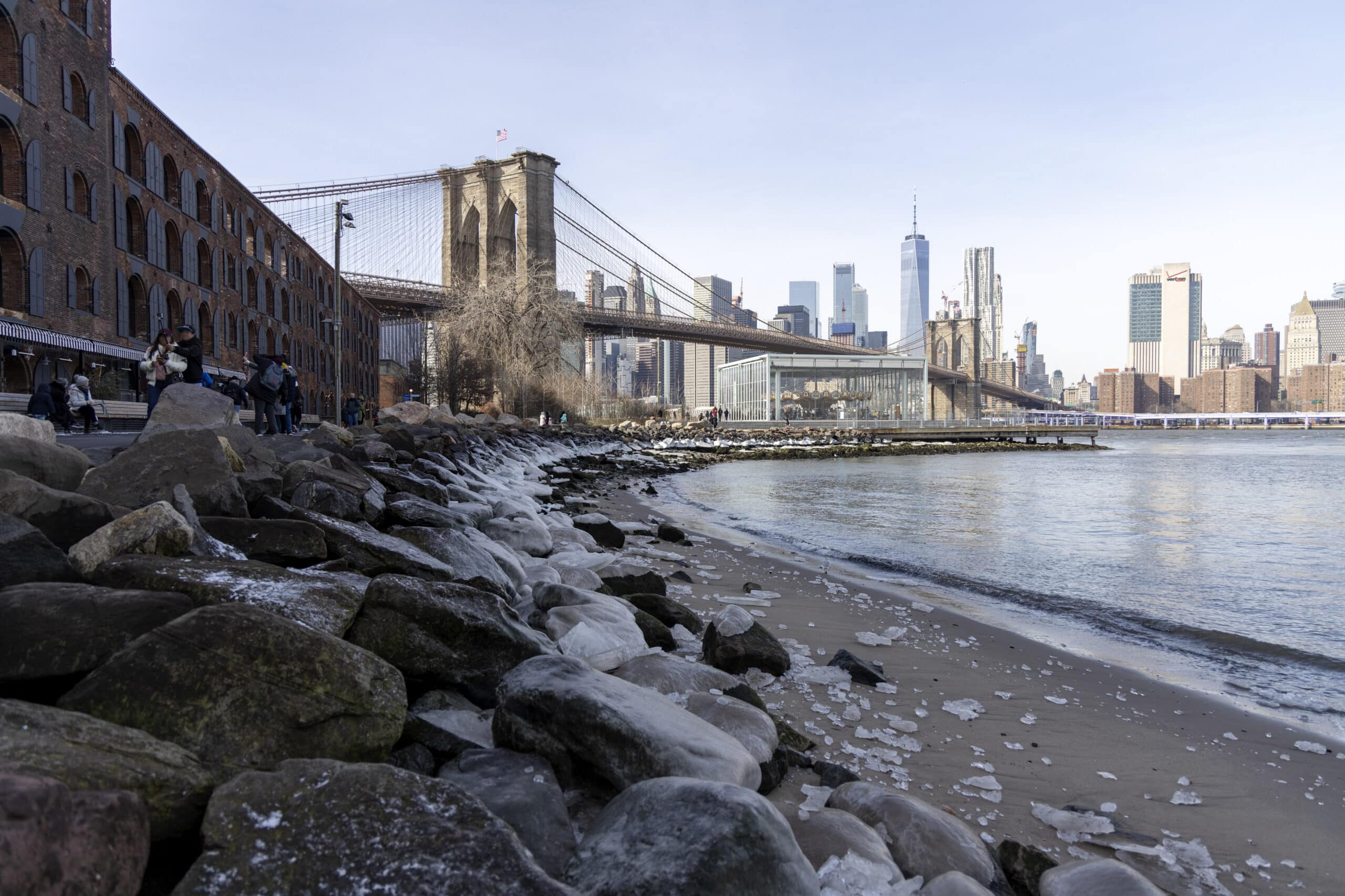 Blick auf die Brooklyn Bridge und die Skyline von Manhattan von einer felsigen Küste im Brooklyn Bridge Park, mit Menschen, die das Flussufer genießen. © Fotografie Tomas Rodriguez