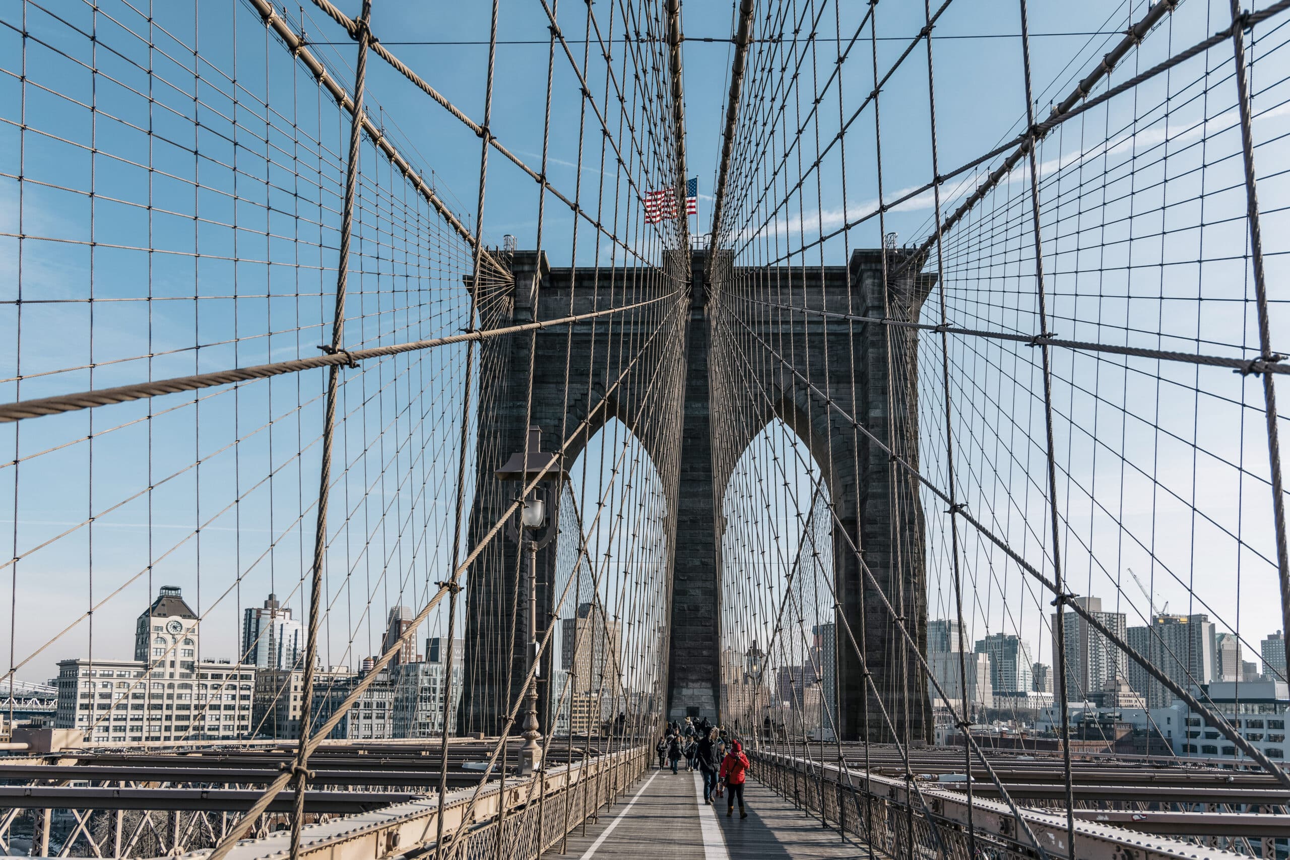 Blick auf die Brooklyn Bridge mit Fußgängern und Radfahrern, mit den ikonischen gotischen Bögen und Kabeldrähten, mit der Skyline von New York im Hintergrund unter einem klaren blauen Himmel. © Fotografie Tomas Rodriguez