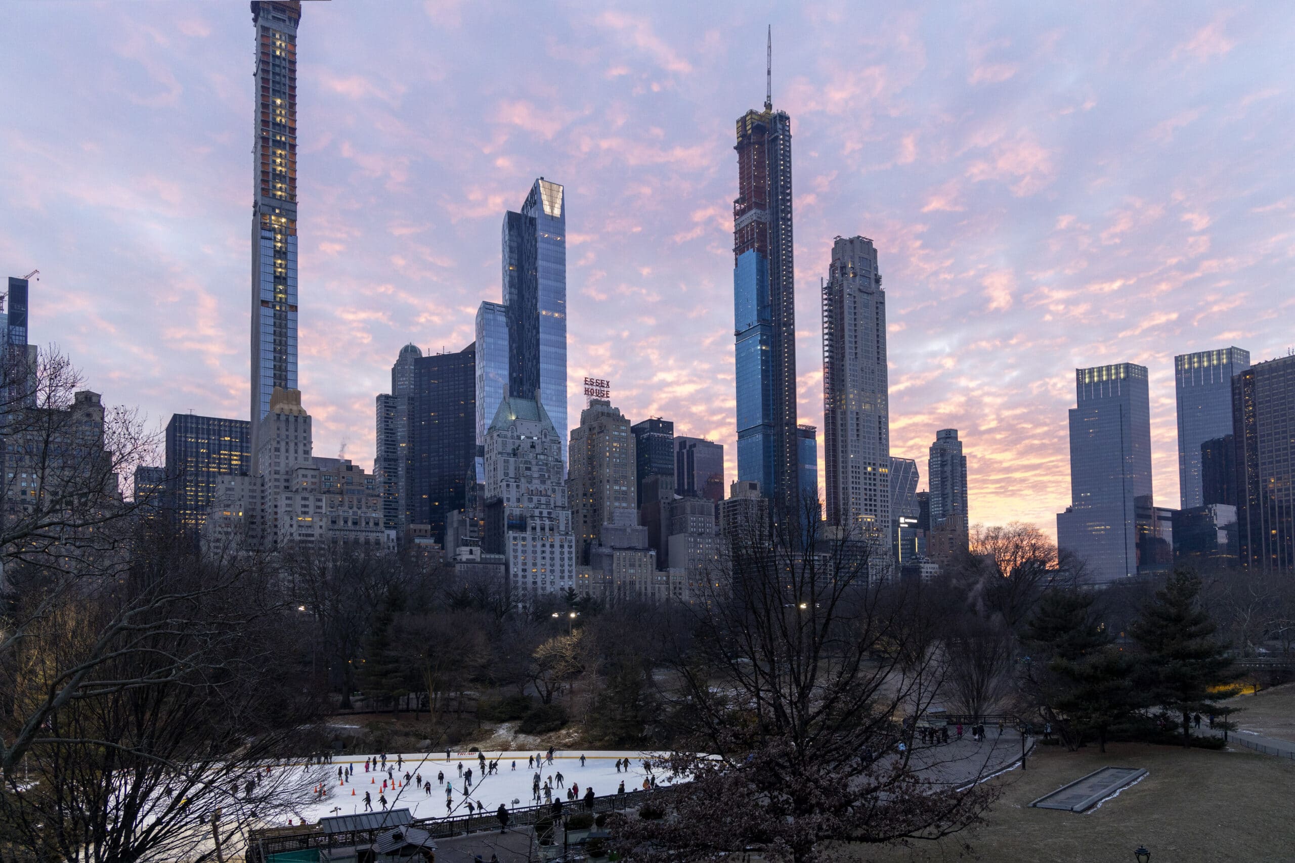 Blick auf eine belebte Eislaufbahn im Central Park in der Abenddämmerung mit hoch aufragenden Wolkenkratzern im Hintergrund und einem leuchtend rosa und violetten Himmel. © Fotografie Tomas Rodriguez