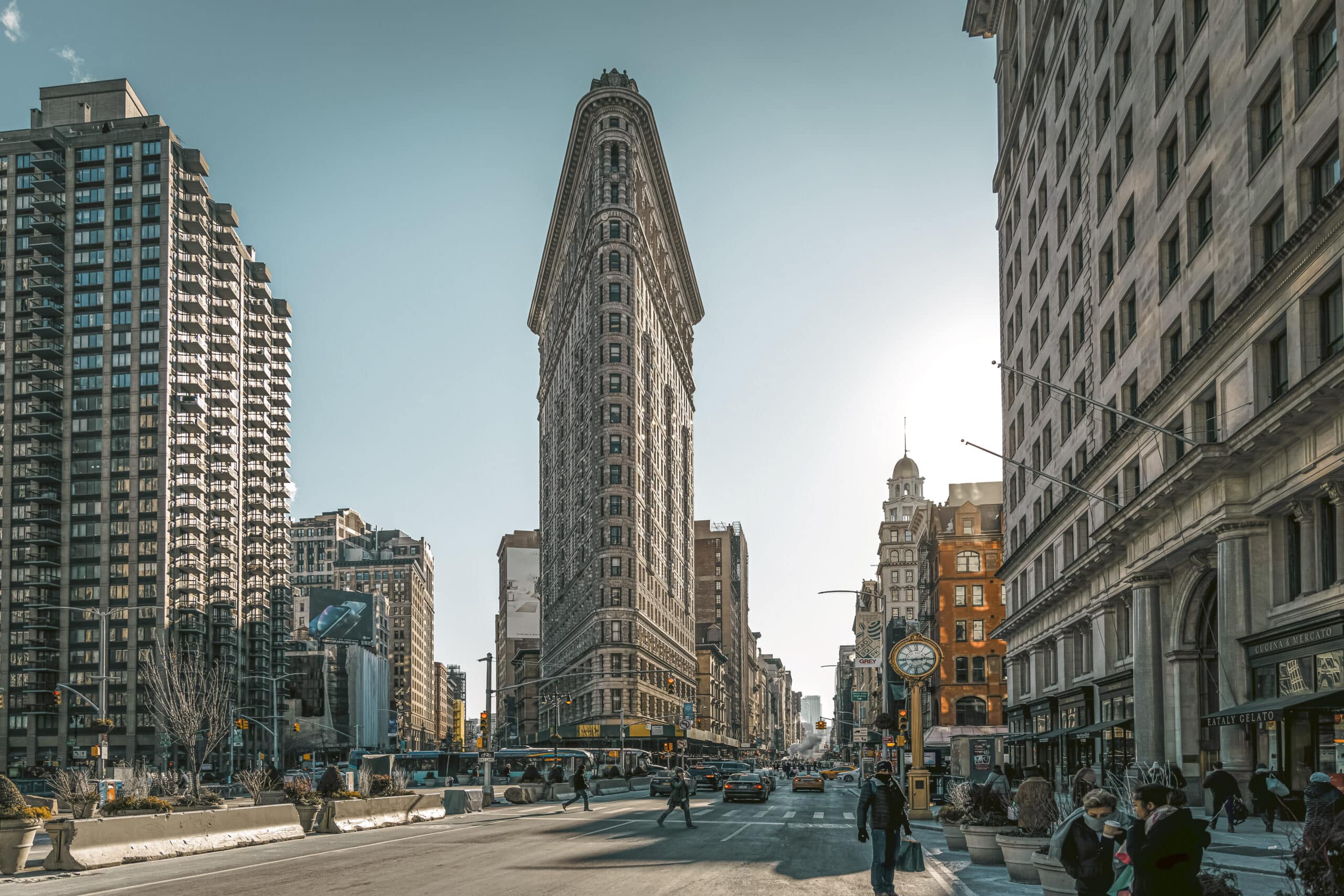 Ein Blick von der Straße auf das ikonische Flatiron Building in New York City an einem sonnigen Tag, mit Fußgängern und Verkehr auf den umliegenden Straßen und anderen modernen und älteren Gebäuden, die das historische Bauwerk flankieren. © Fotografie Tomas Rodriguez