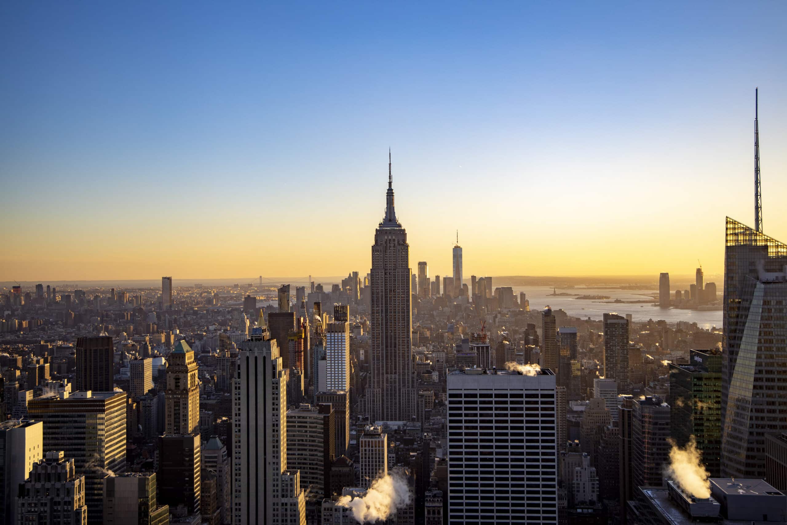 Ein malerischer Blick bei Sonnenaufgang über der Skyline von New York City mit dem Empire State Building im Mittelpunkt, wobei helles Sonnenlicht einen warmen Glanz über die Stadtlandschaft wirft. © Fotografie Tomas Rodriguez