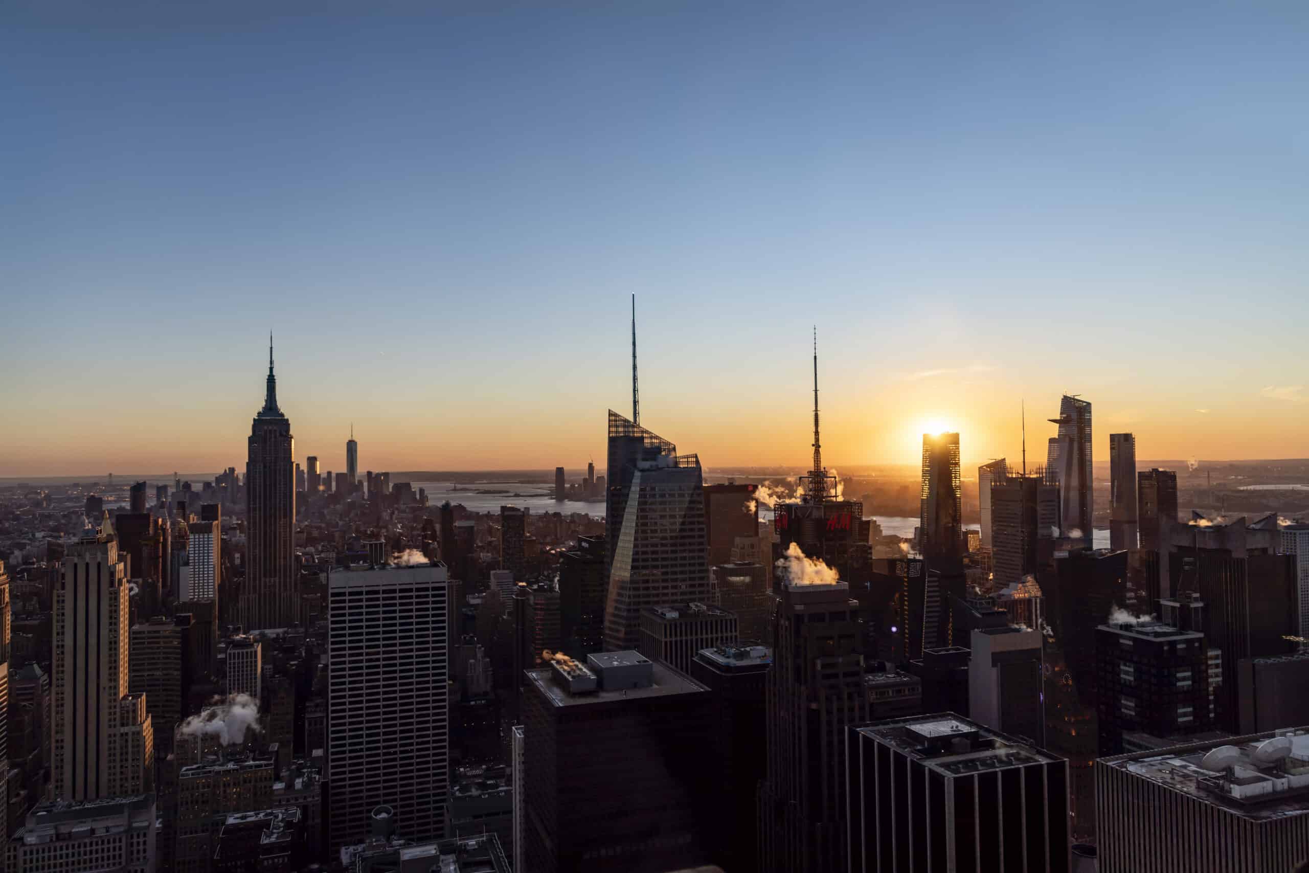 Blick auf die Skyline einer Stadt bei Sonnenuntergang. Hervorzuheben sind markante Wolkenkratzer mit einem warmen Glanz am Horizont unter einem klaren Himmel. © Fotografie Tomas Rodriguez