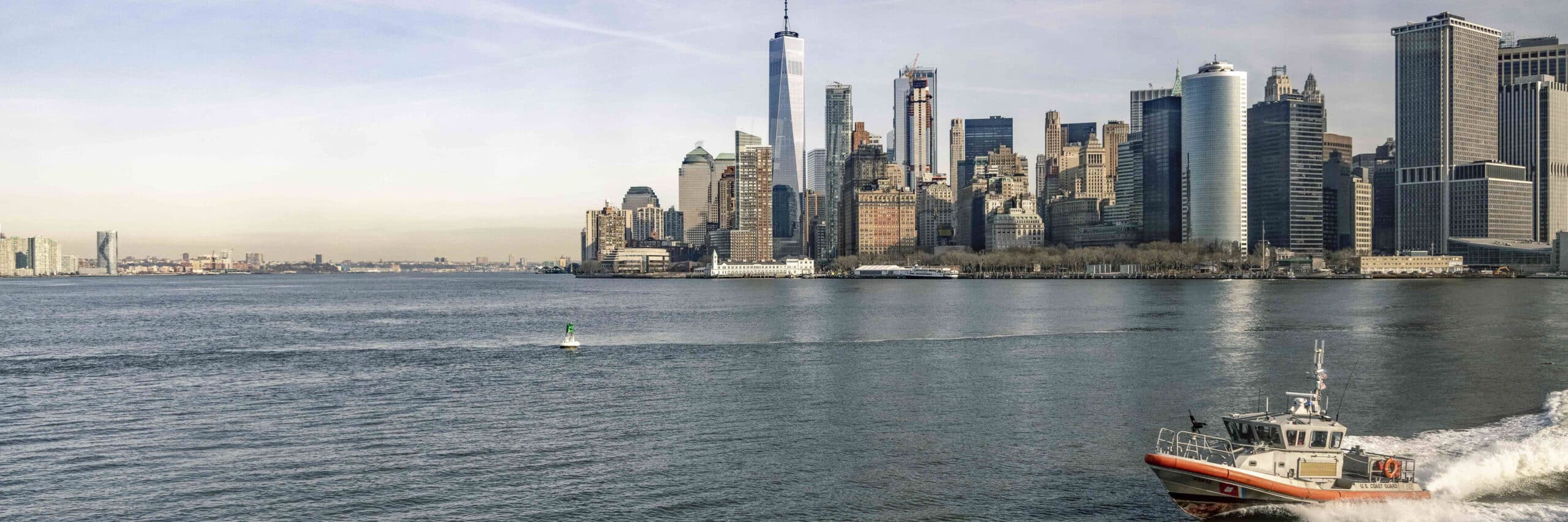 Ein Panoramablick auf die Skyline von Lower Manhattan über einen Fluss hinweg, mit hohen Wolkenkratzern, darunter das One World Trade Center. Im Vordergrund rast ein Boot der Küstenwache über das Wasser. © Fotografie Tomas Rodriguez