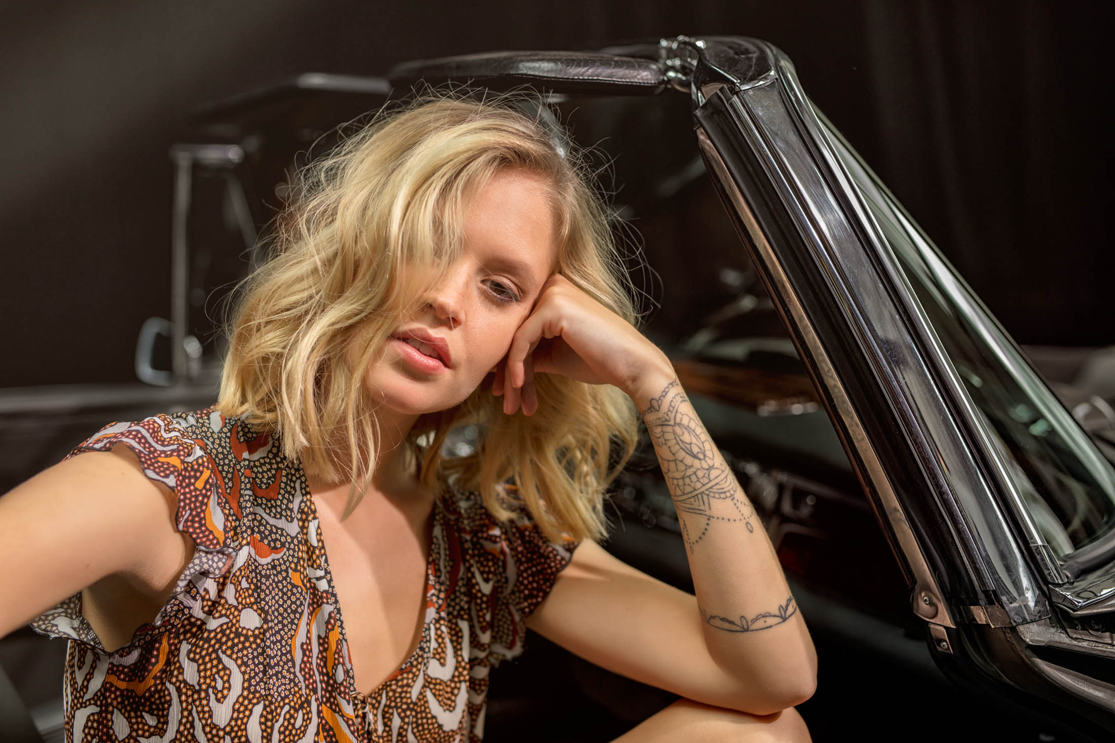 Eine Frau mit blonden Haaren und einer Tätowierung auf dem Arm lehnt nachdenklich an einem Oldtimer, den Kopf auf der Hand abgestützt, im Hintergrund ist unscharf ein Schlagzeug zu sehen. © Fotografie Tomas Rodriguez