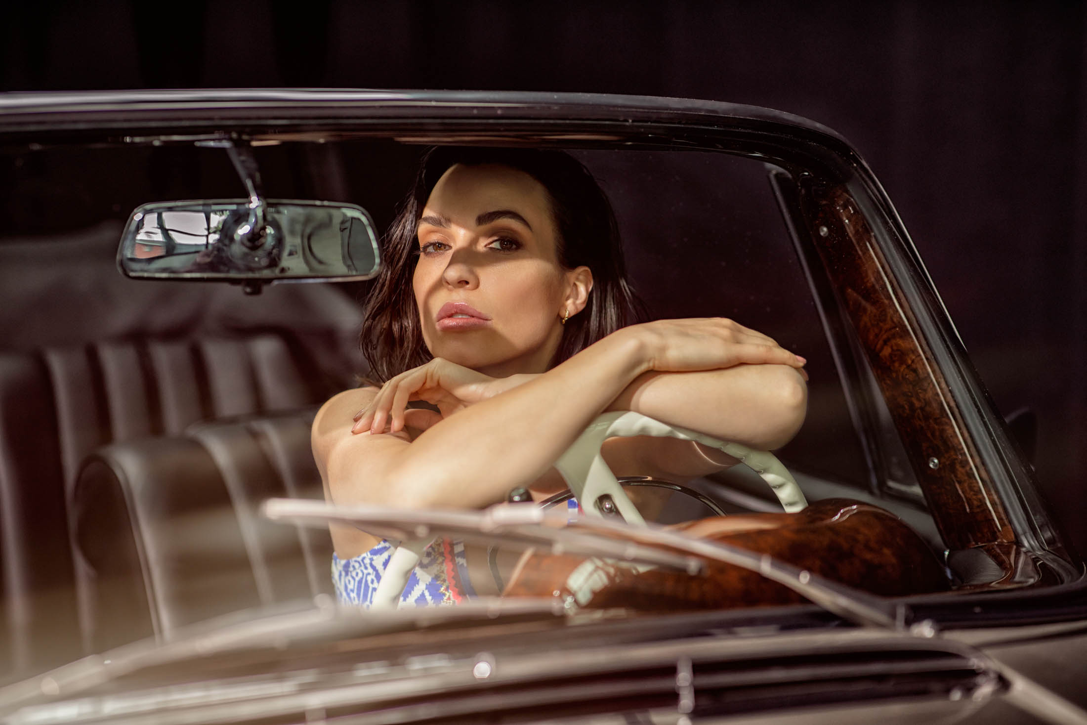 Eine Frau mit dunklem Haar blickt mit nachdenklichem Gesichtsausdruck durch das offene Fenster eines Oldtimers, stützt ihre Arme und ihr Kinn auf die Tür. © Fotografie Tomas Rodriguez