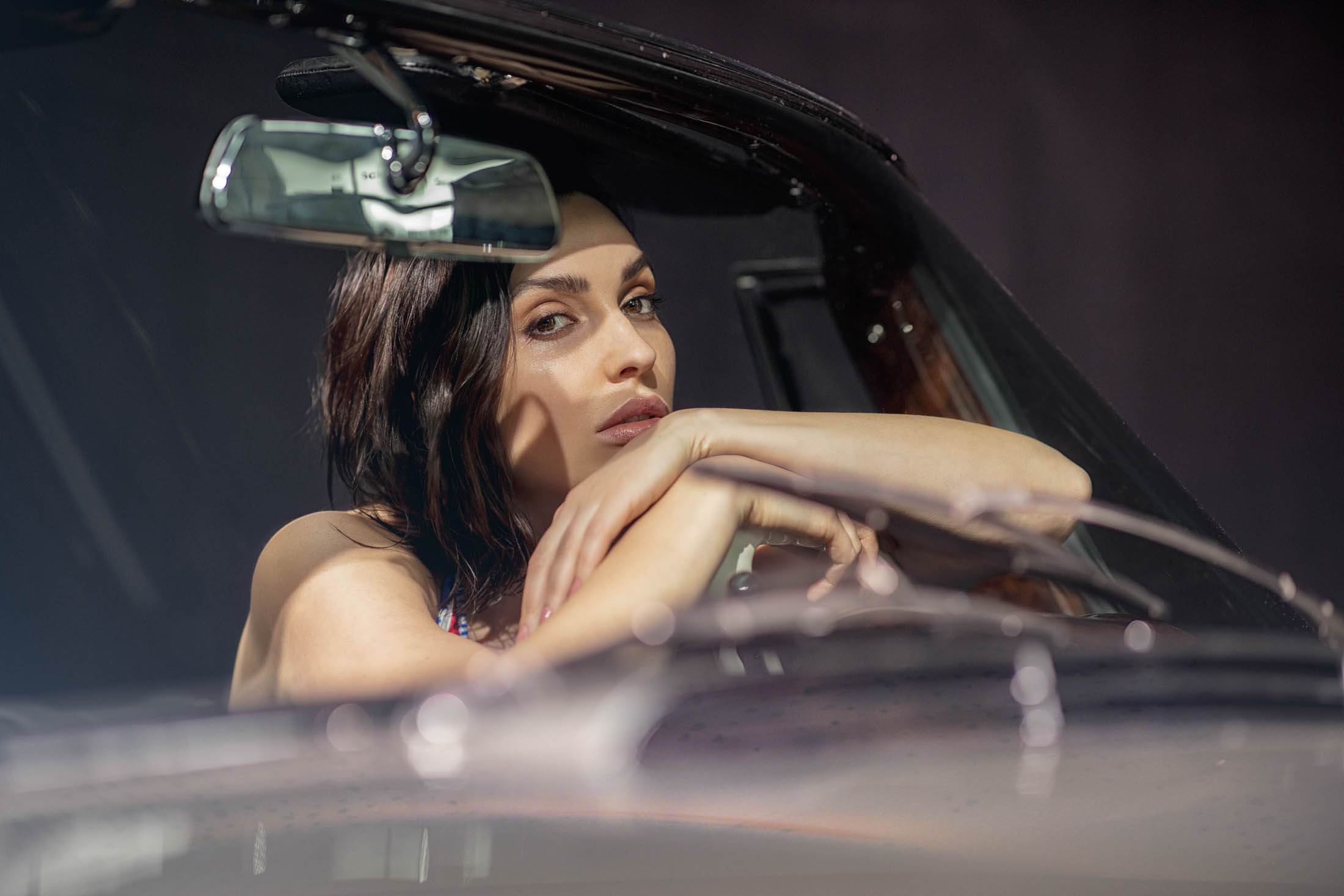 Eine Frau mit dunklem Haar stützt ihr Kinn auf ihre Hände, während sie auf dem Fahrersitz eines Autos sitzt und mit nachdenklichem Gesichtsausdruck durch die Windschutzscheibe blickt. © Fotografie Tomas Rodriguez