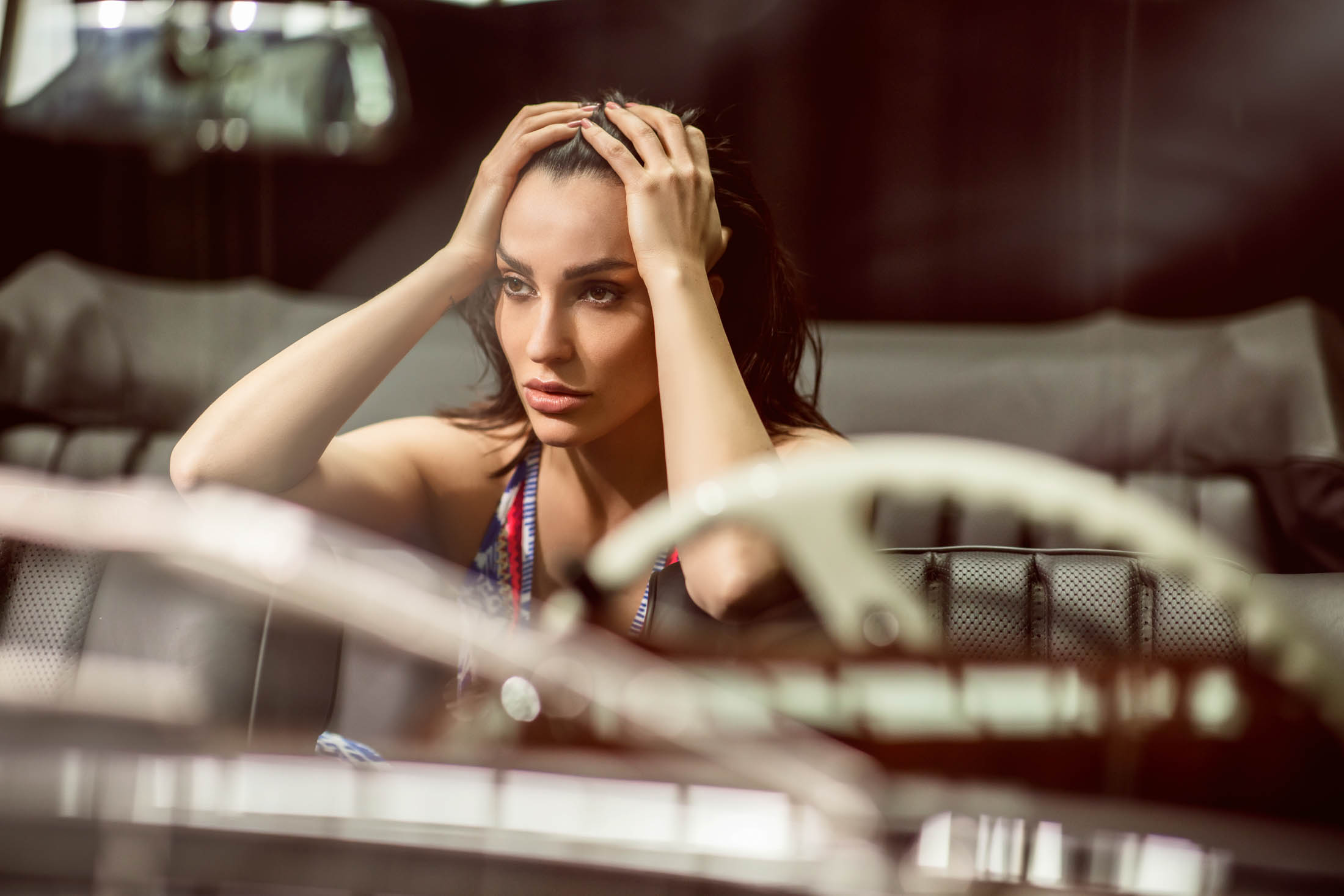 Eine Frau mit nachdenklichem Gesichtsausdruck sitzt in einem Auto und hat die Hände auf dem Kopf abgelegt. Im Vordergrund sind Lenkrad und Armaturenbrett zu sehen, die den Innenraum eines modernen Fahrzeugs widerspiegeln. © Fotografie Tomas Rodriguez