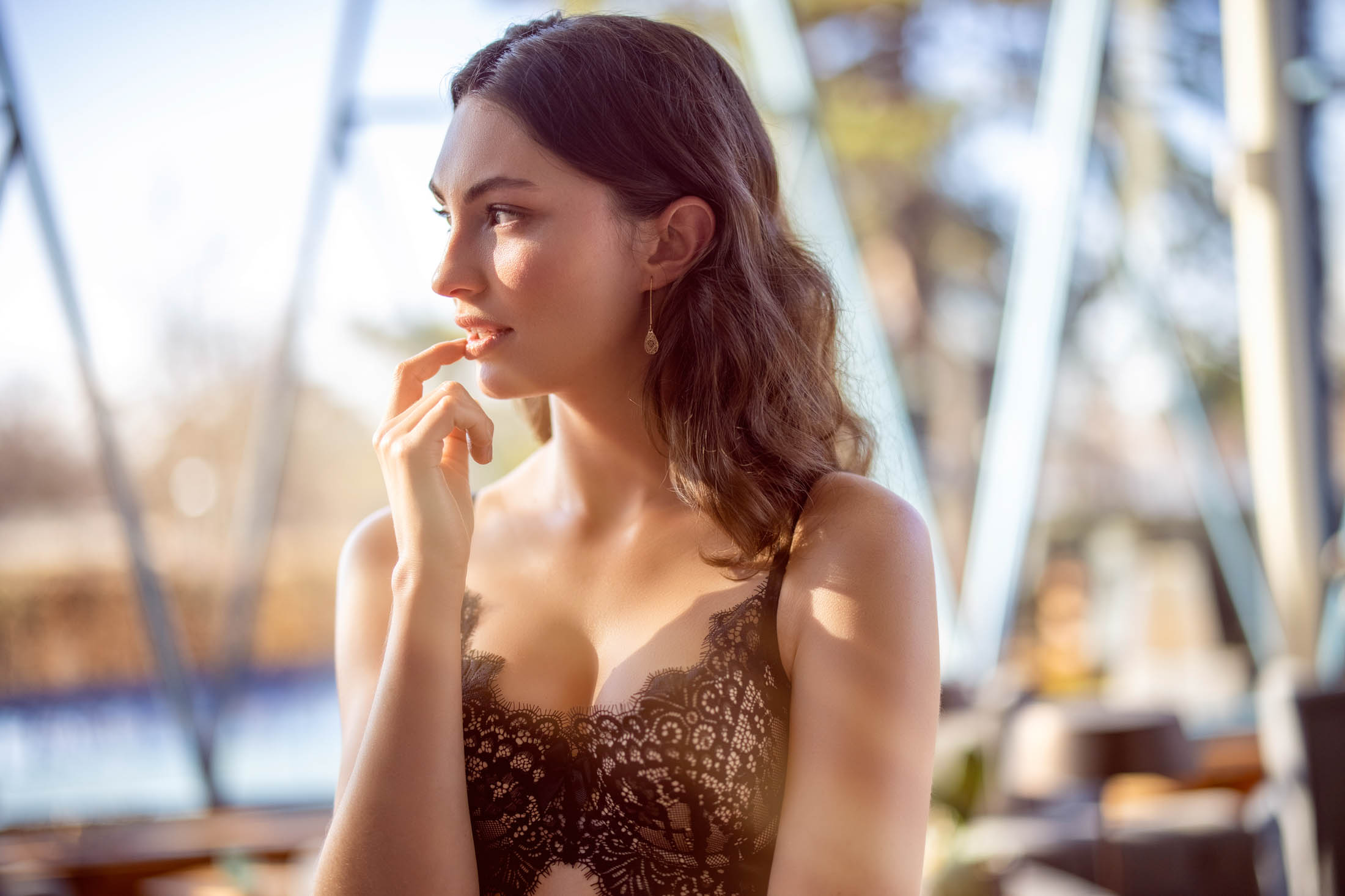 Eine junge Frau in einem schwarzen Spitzenkleid sitzt nachdenklich da, die Finger auf dem Kinn, und blickt in die Ferne, vor dem sanft beleuchteten, verschwommenen Hintergrund einer sonnigen Außenlandschaft. © Fotografie Tomas Rodriguez