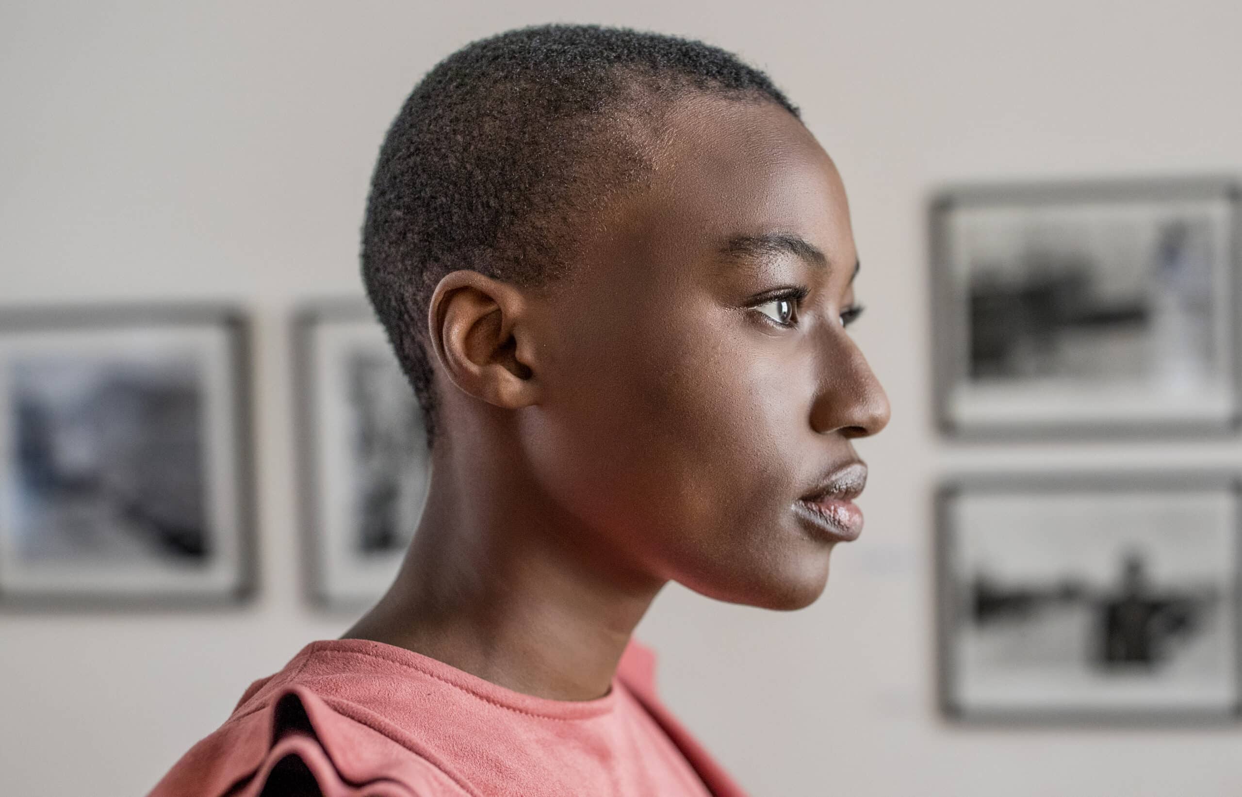 Profilansicht einer jungen Person mit kurzen Haaren, die ein rosa T-Shirt trägt und nachdenklich zur Seite blickt. Im Hintergrund sind gerahmte Graustufenfotos auf einer hellen Wand zu sehen. © Fotografie Tomas Rodriguez