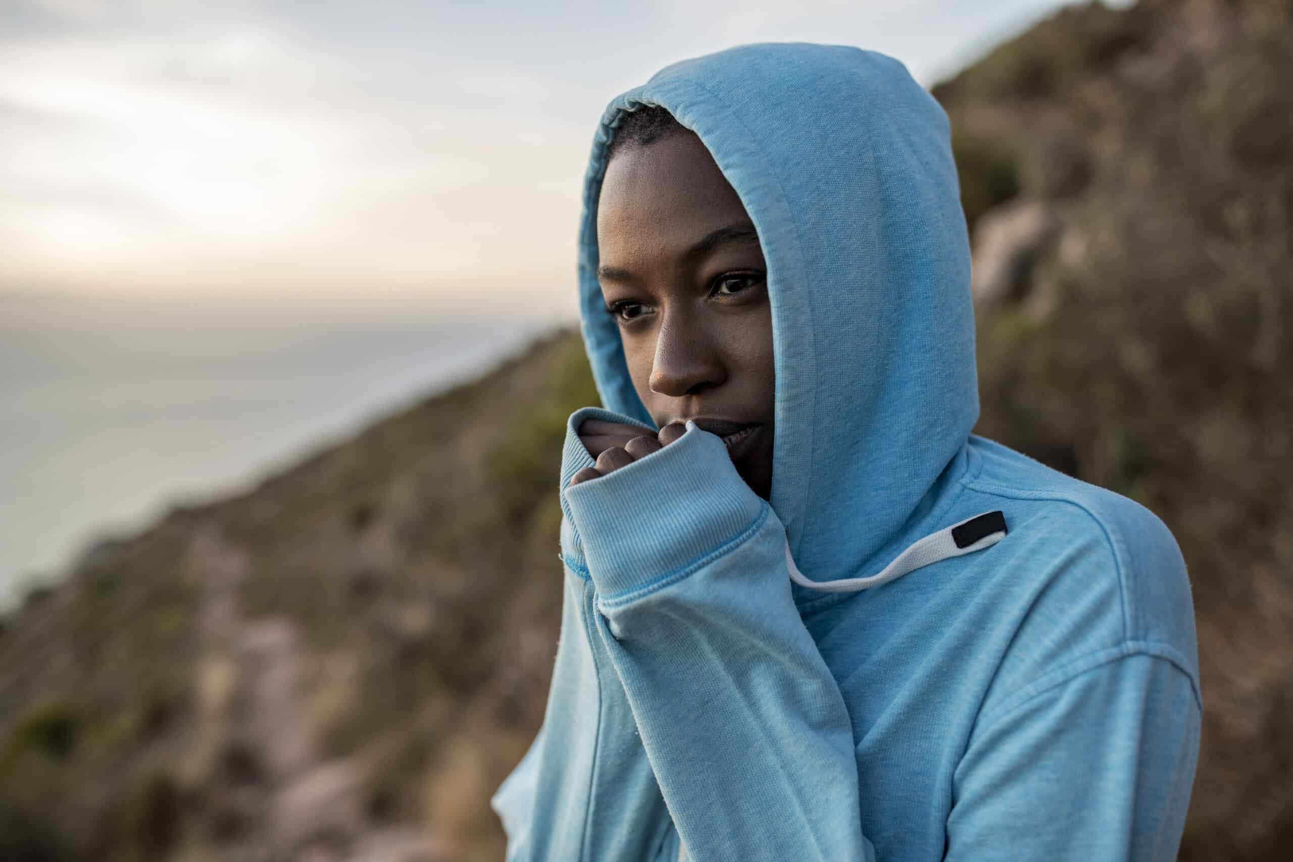 Eine nachdenkliche junge Frau in einem blauen Kapuzenpullover steht im Freien. Ihr Gesicht ist teilweise von der Kapuze bedeckt. Im Hintergrund ist eine heitere Landschaft mit weichem Fokus zu sehen. © Fotografie Tomas Rodriguez
