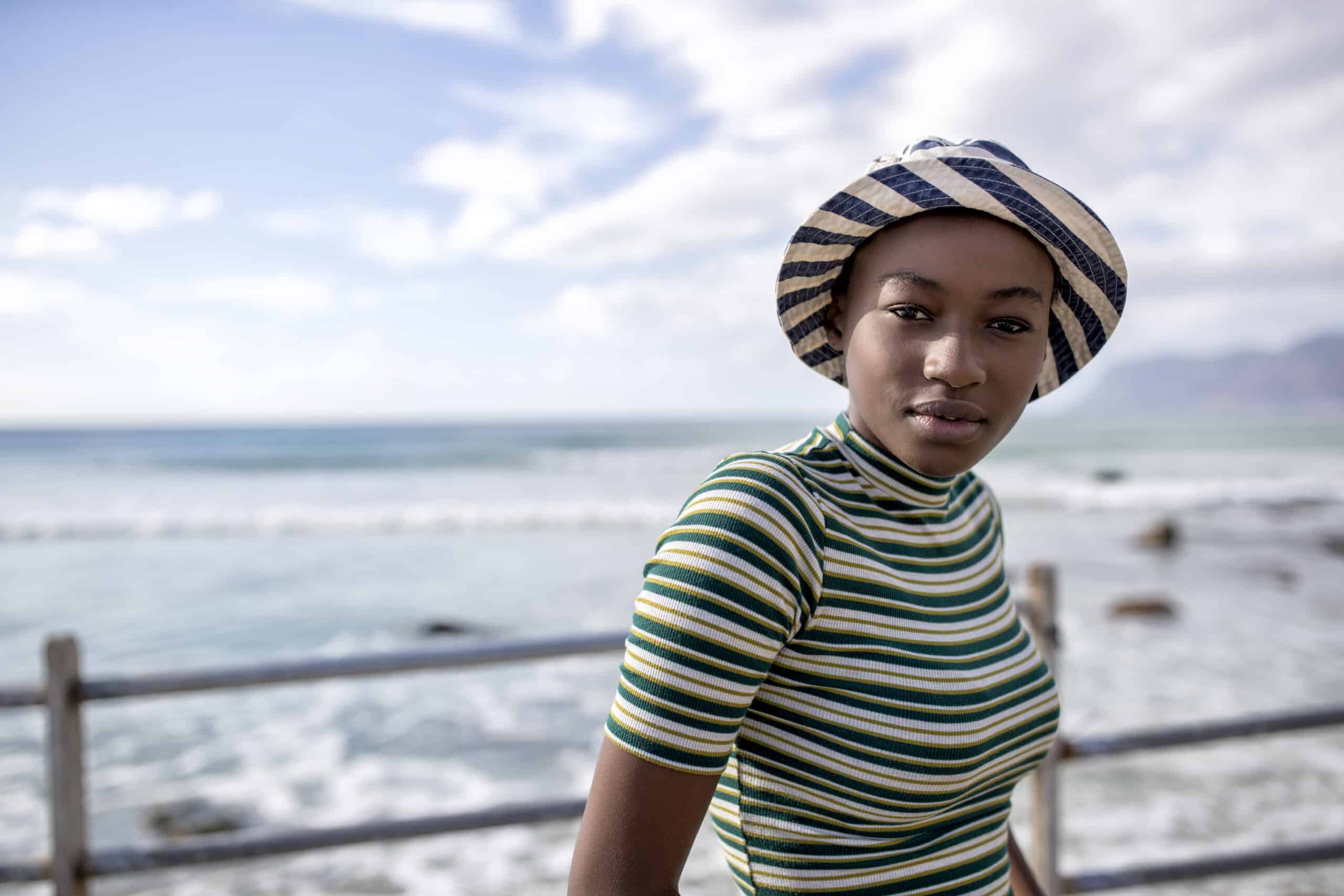 Eine junge Frau mit gestreiftem Hut und Hemd steht an einem Geländer am Meer, im Hintergrund sind das ruhige Meer und die Berge zu sehen. © Fotografie Tomas Rodriguez