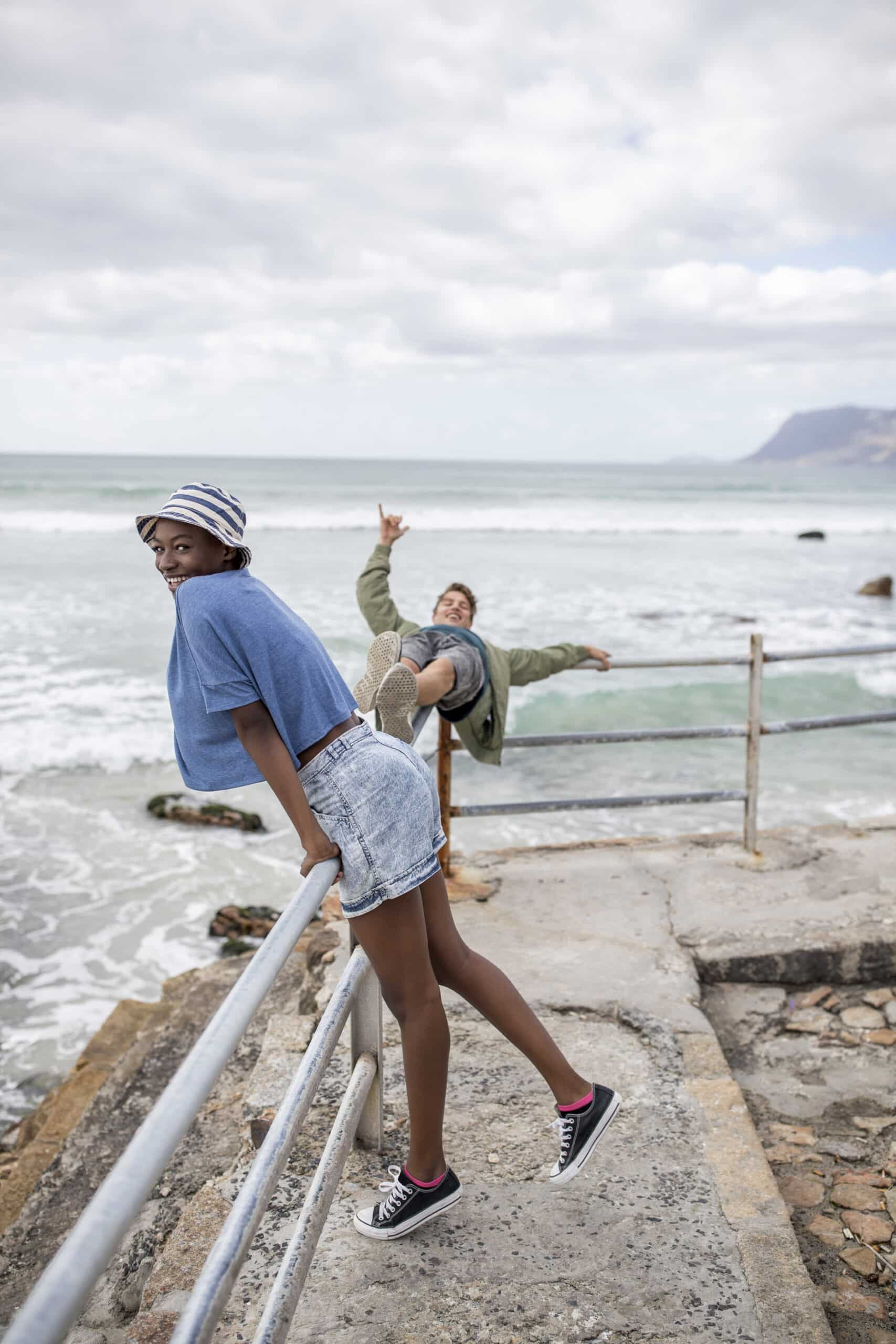 Eine fröhliche junge Frau in Jeanskleidung und Beanie steht am Geländer am Meer und lächelt in die Kamera, während im Hintergrund eine andere Person verspielt mit ausgestreckten Armen posiert und die Meereswellen brechen. © Fotografie Tomas Rodriguez