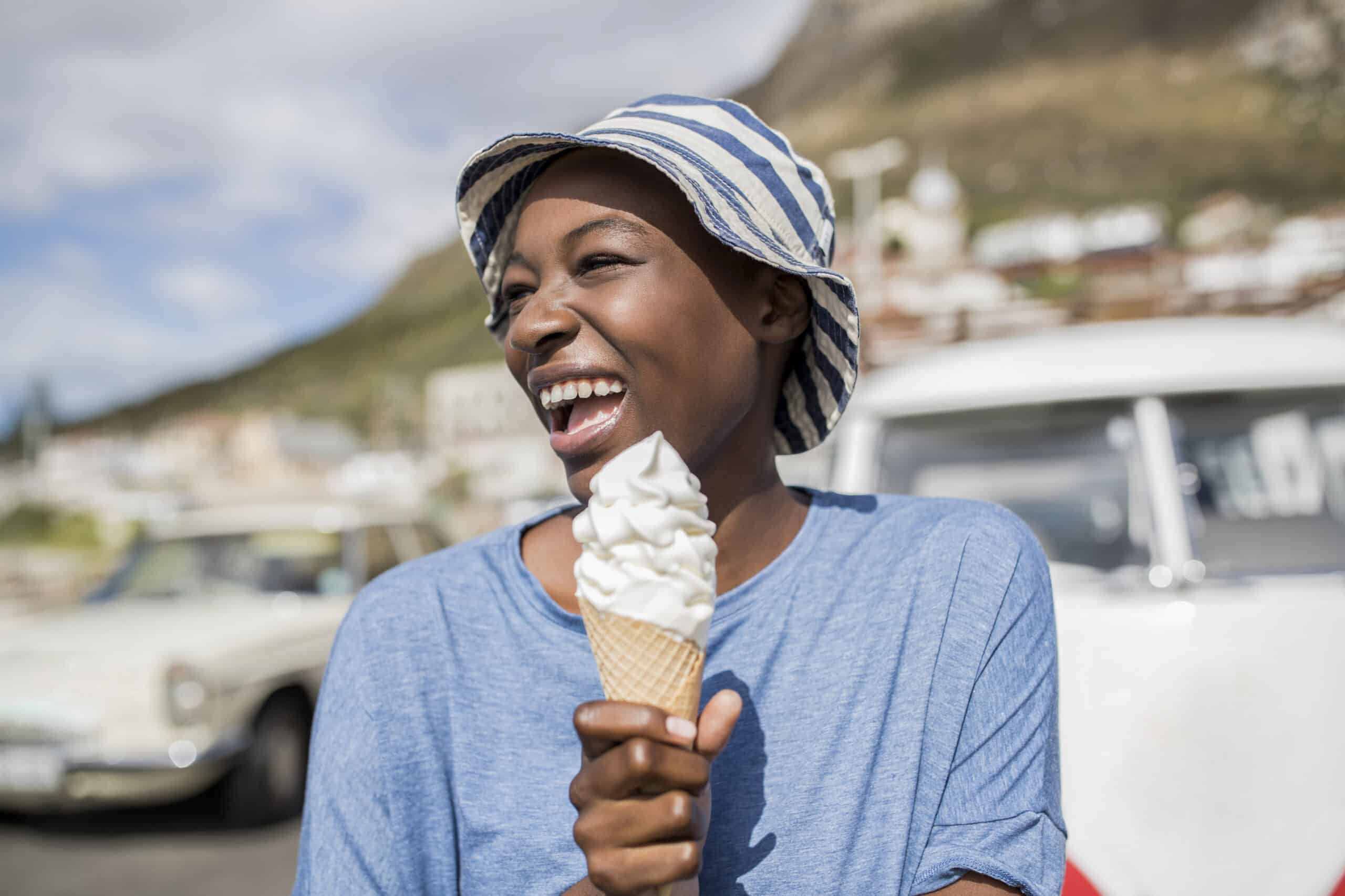 Eine fröhliche Frau mit gestreiftem Hut genießt an einem sonnigen Tag eine Eistüte, im Hintergrund sind Oldtimer und ein Berg zu sehen. © Fotografie Tomas Rodriguez